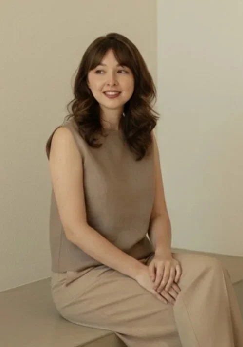 A young woman with shoulder-length brown hair and a warm smile, sitting on a bench against a plain light-colored wall, wearing a sleeveless beige top and matching wide-leg pants.