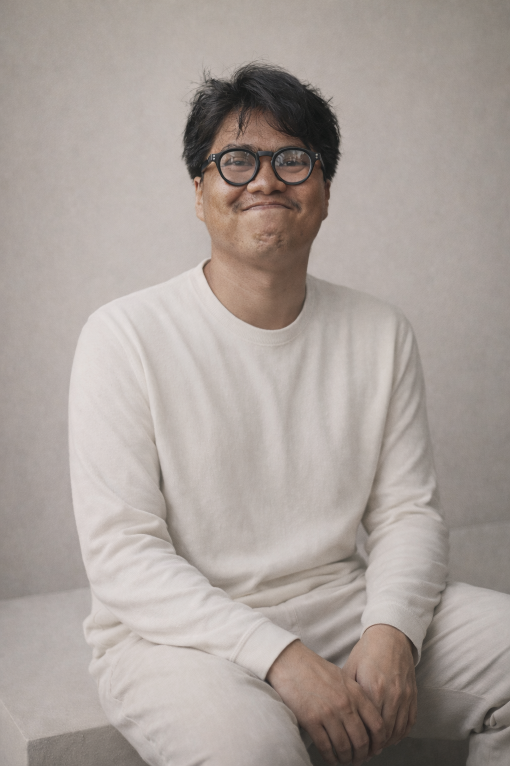 A man with glasses wearing a white long sleeve shirt and white pants sitting against a plain background, smiling.