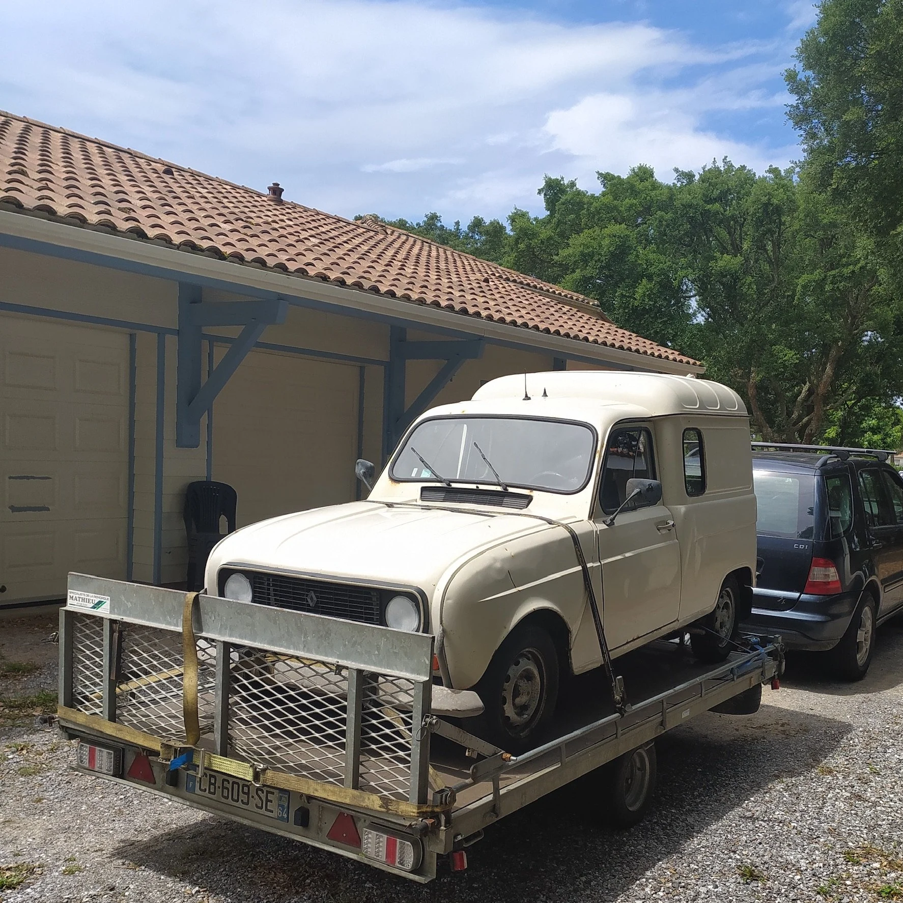 Une petite voiture blanche ancienne est placée sur un plateau de remorque. Elle est attachée avec une sangle. Il y a une maison avec un toit en tuiles en arrière-plan et une voiture noire garée à côté.