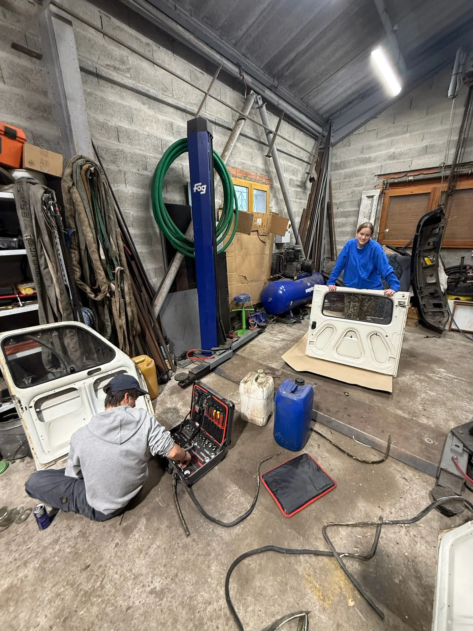 Deux personnes travaillent dans un atelier de réparation de voitures. Un homme est assis sur le sol, manipulant un outil électrique, et une femme en haut d'une caisse vitrée de voiture, souriante, portant une veste bleue.