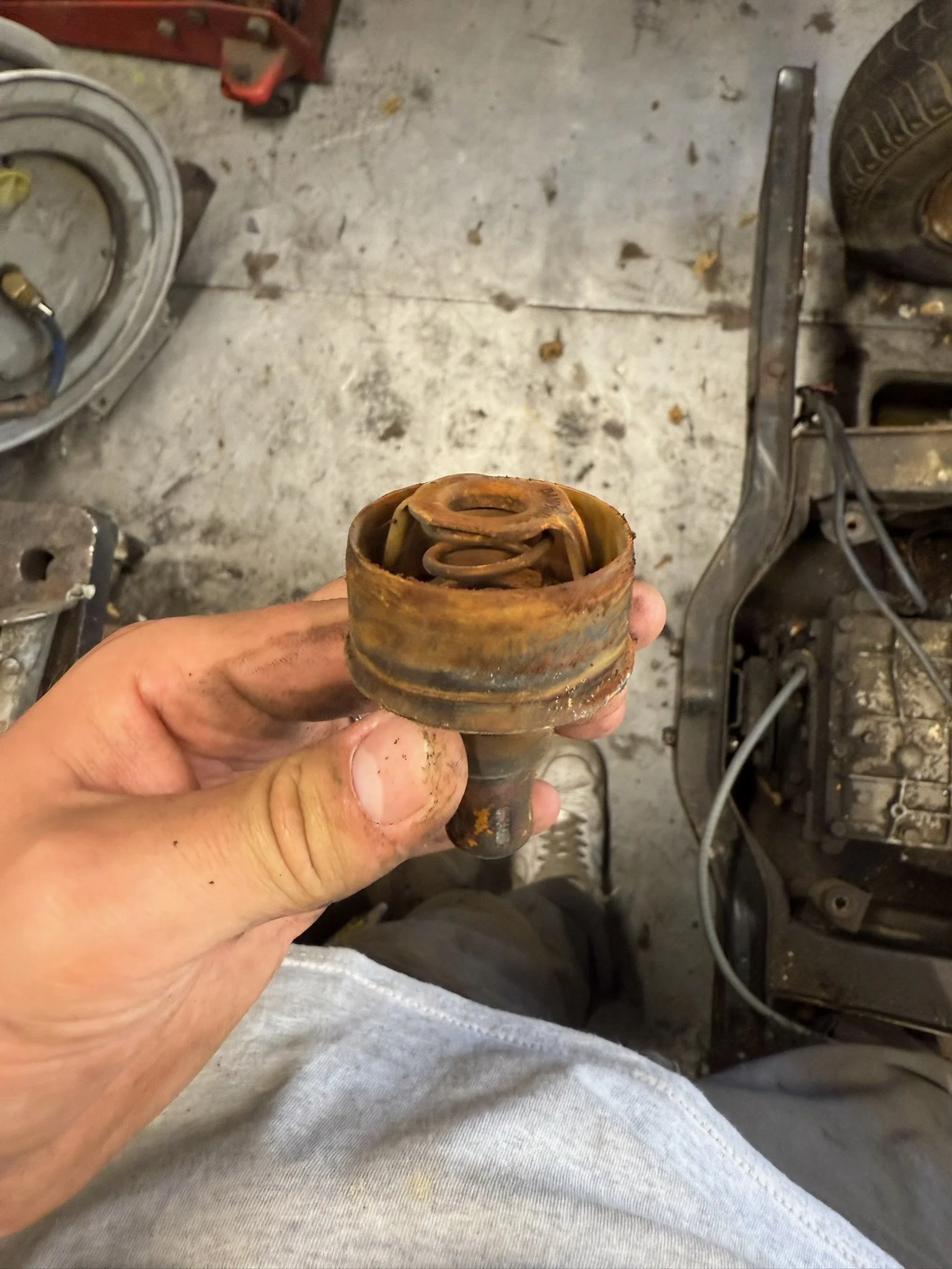 Main tenant une pièce mécanique rouillée, probablement une pièce de moteur ou suspension d'une voiture, dans un atelier ou garage avec un sol en béton usé.