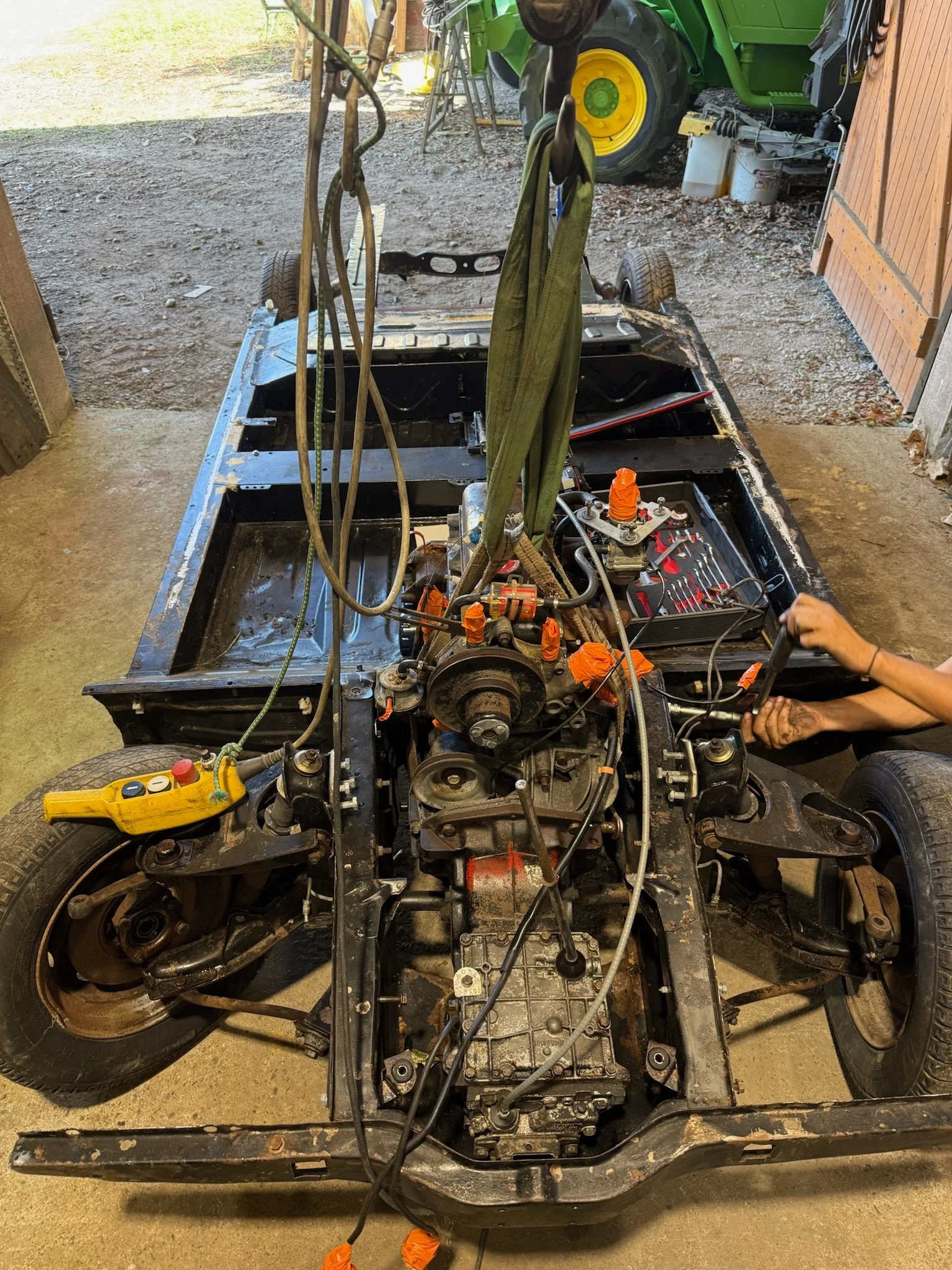 Un châssis de voiture sans carrosserie, en cours de réparation ou de modification, dans un garage. Des outils et des câbles sont visibles, ainsi qu'une personne travaillant sur le véhicule.