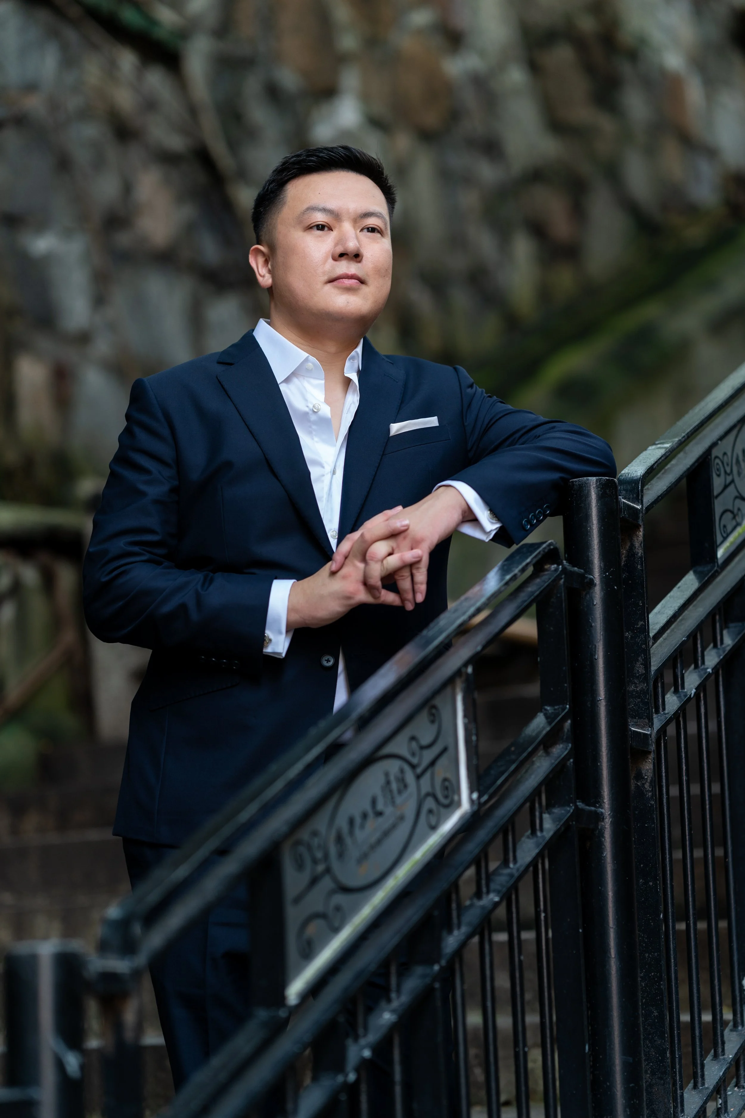 A man in a navy blue suit with a white shirt standing outdoors beside a black metal railing, with a rocky background.