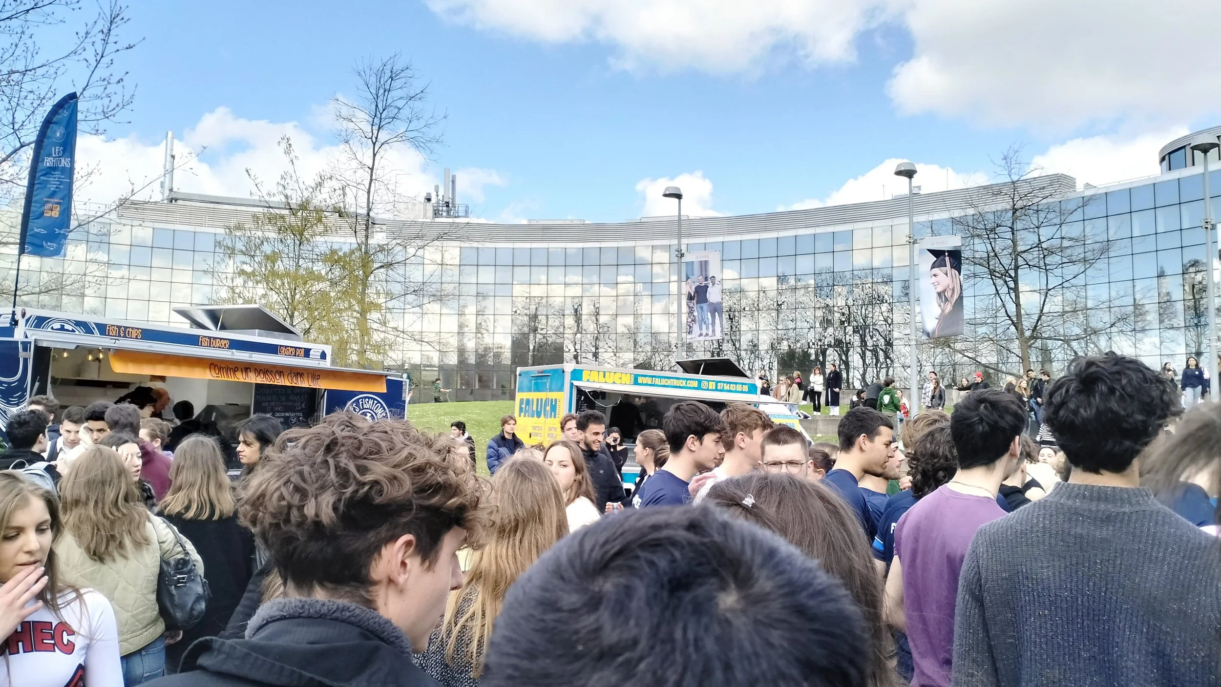 Une foule de jeunes adultes se rassemble à l'extérieur devant un bâtiment en verre, avec le food truck faluch'truck et un ciel bleu avec des nuages.