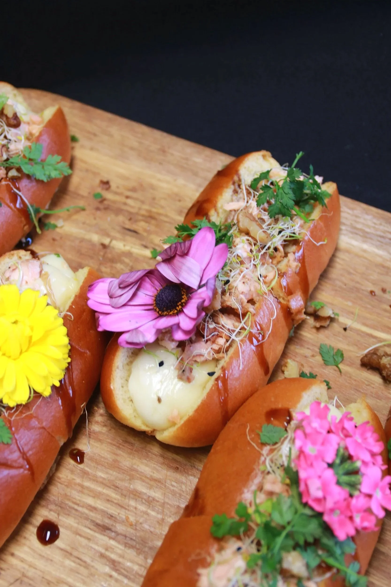 Sandwichs dans des petits pains garnis de fromage fondu, de fines herbes, de germes, de sauce et décorés avec des fleurs comestibles colorées sur une planche en bois.