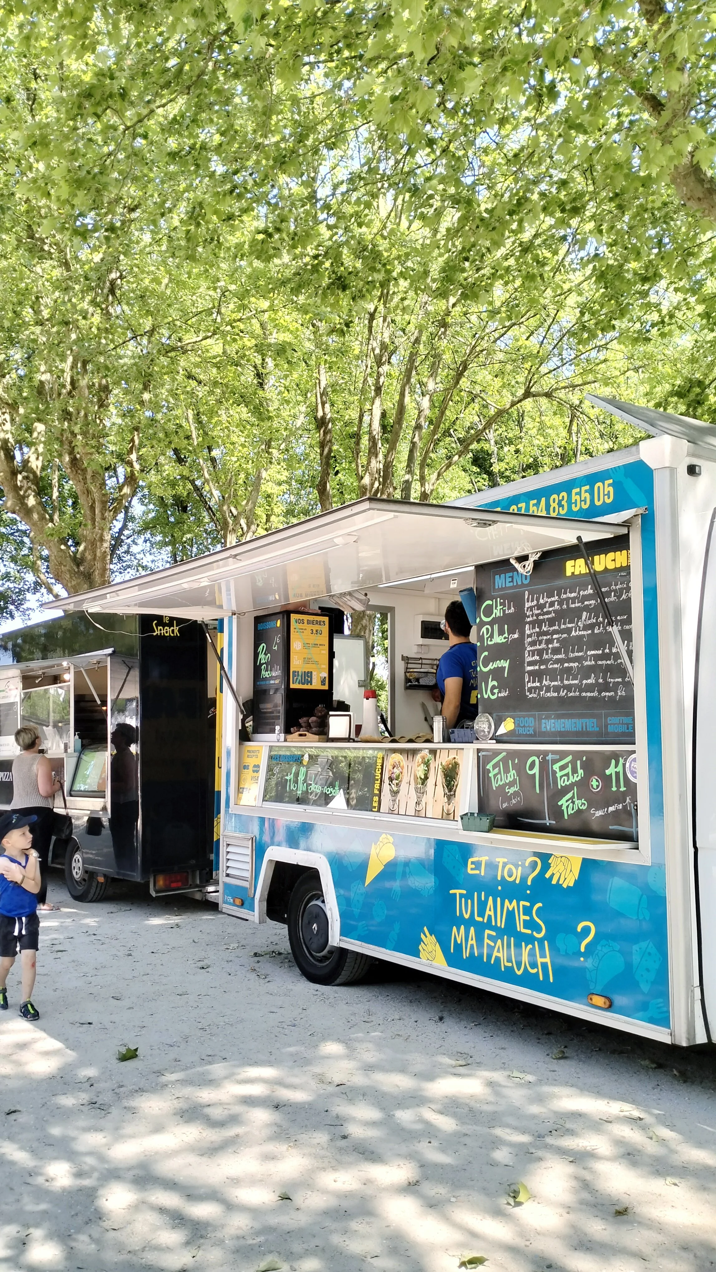 Le camion de Faluch'Truck avec un menu écrit à la craie, un arbre vert en arrière-plan et des personnes qui attendent ou se trouvent à proximité.