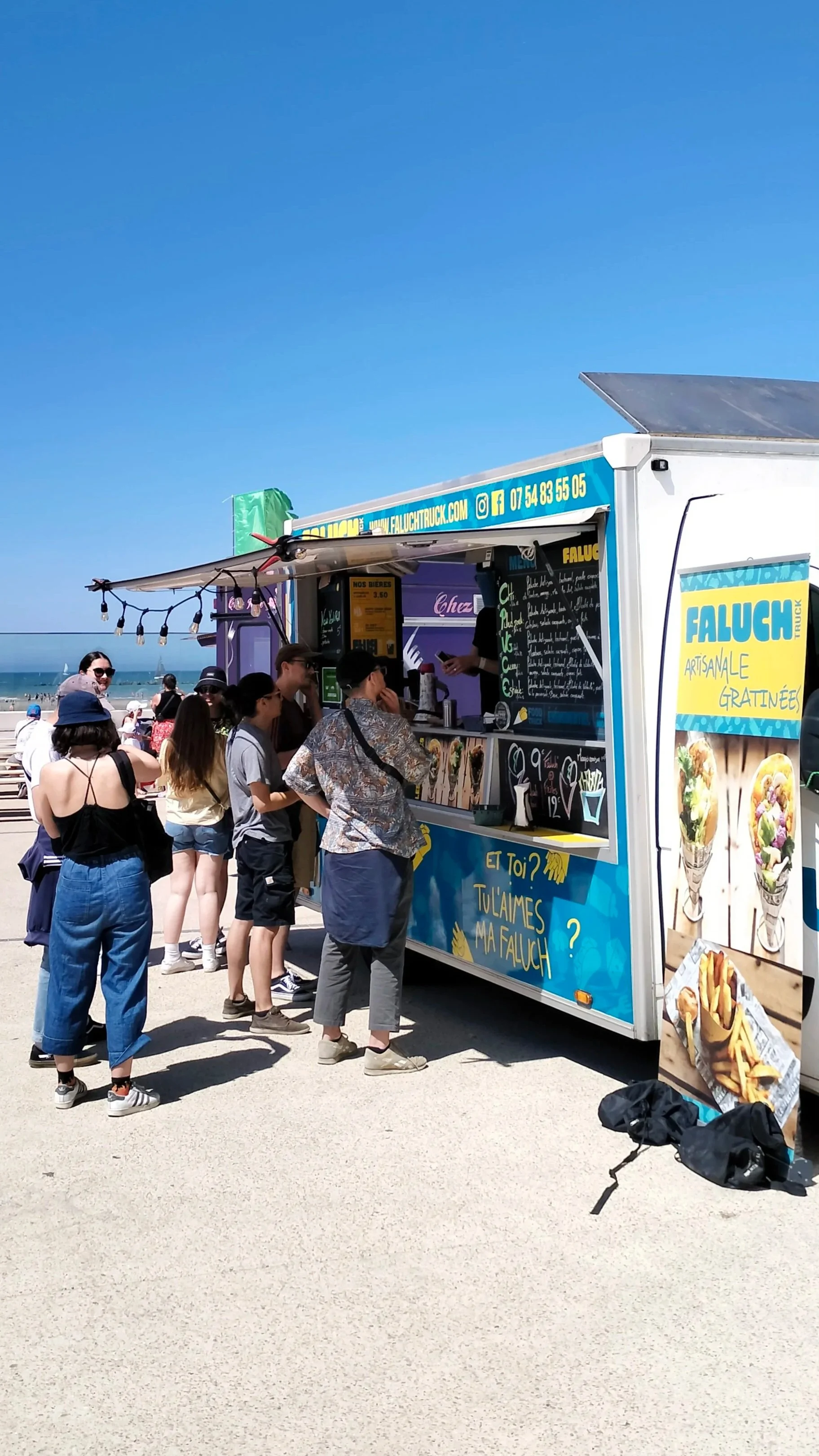 Une file d'attente devant le food truck Faluch'truck en bord de plage, avec des personnes faisant la queue pour commander des crêpes garnies, avec un ciel bleu clair et la mer en arrière-plan.