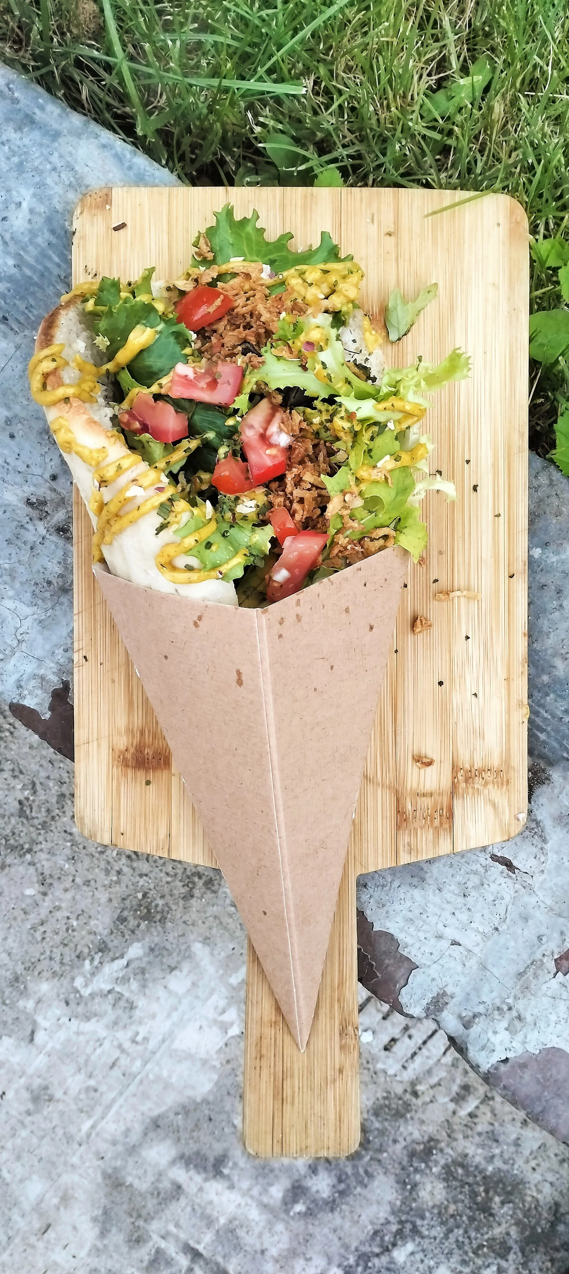 Un taco garni de viande, salade, tomates, moutarde, dans une caissette en papier, posé sur une planche en bois, à l'extérieur.