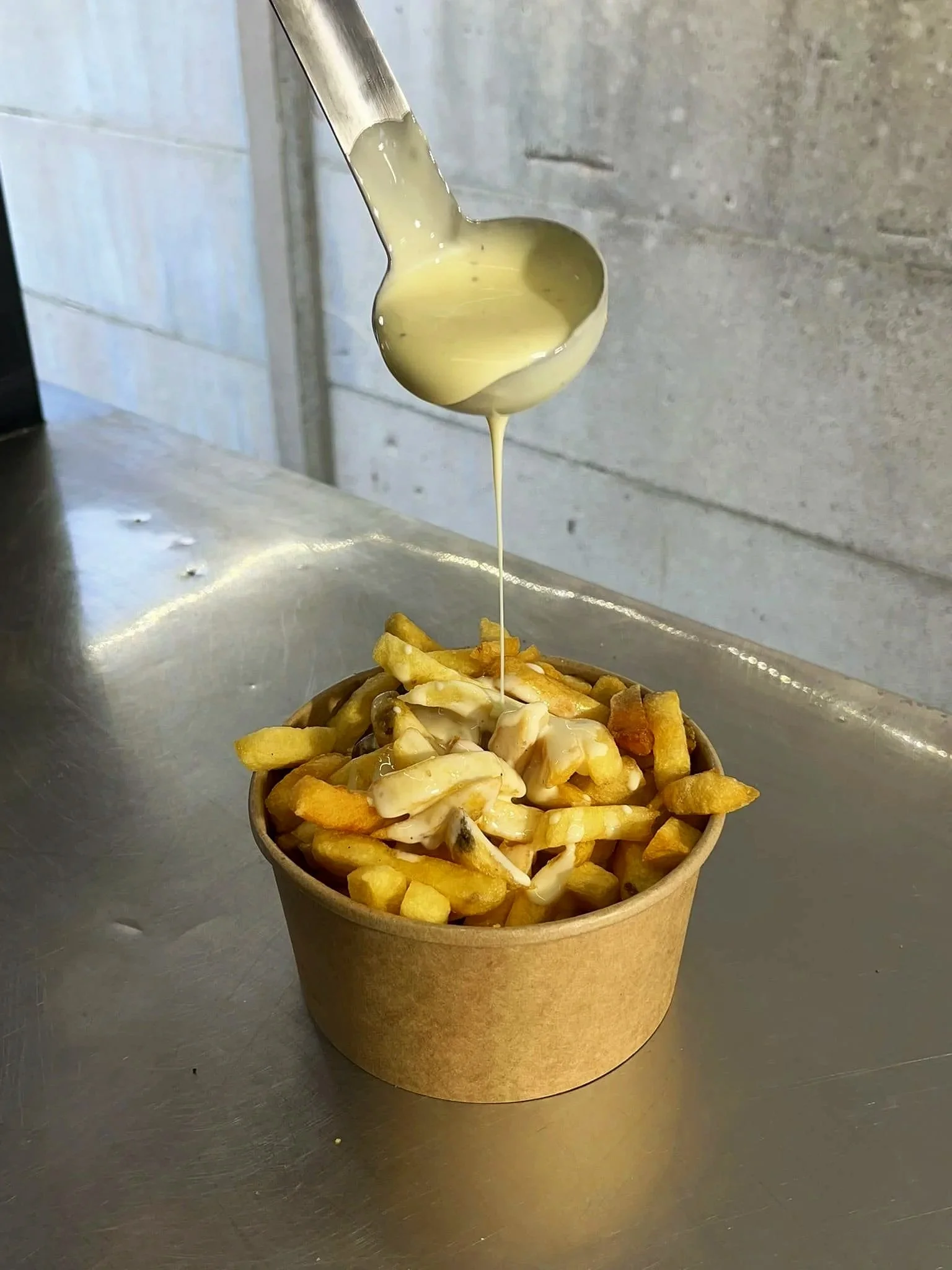 Une cuillère verse une sauce maroilles sur des frites dans un bol en carton.