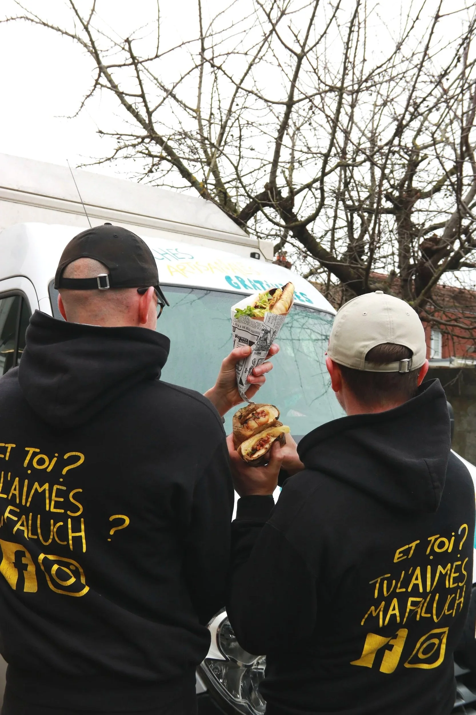 Deux hommes portant des sweats à capuche noirs, ils se tiennent devant le food truck, l'un tient une faluche dans une feuille de papier journal et l'autre une autre partie de la faluche