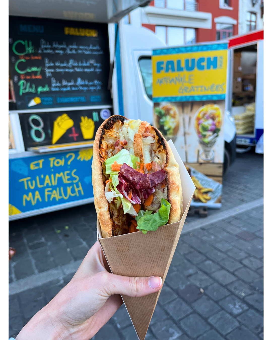 Faluche avec légumes, dans une main, devant un food truck avec panneau coloré en arrière-plan.