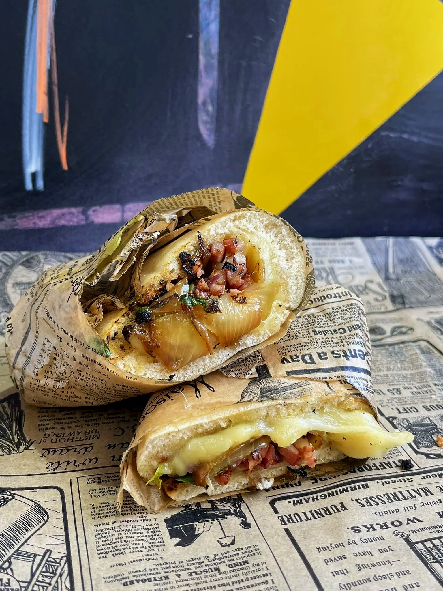 Faluche avec du fromage fondu, du jambon, des oignons grillés, et des herbes, enveloppé dans du papier à motif journal, posé sur une surface recouverte de papier semblable à un journal.