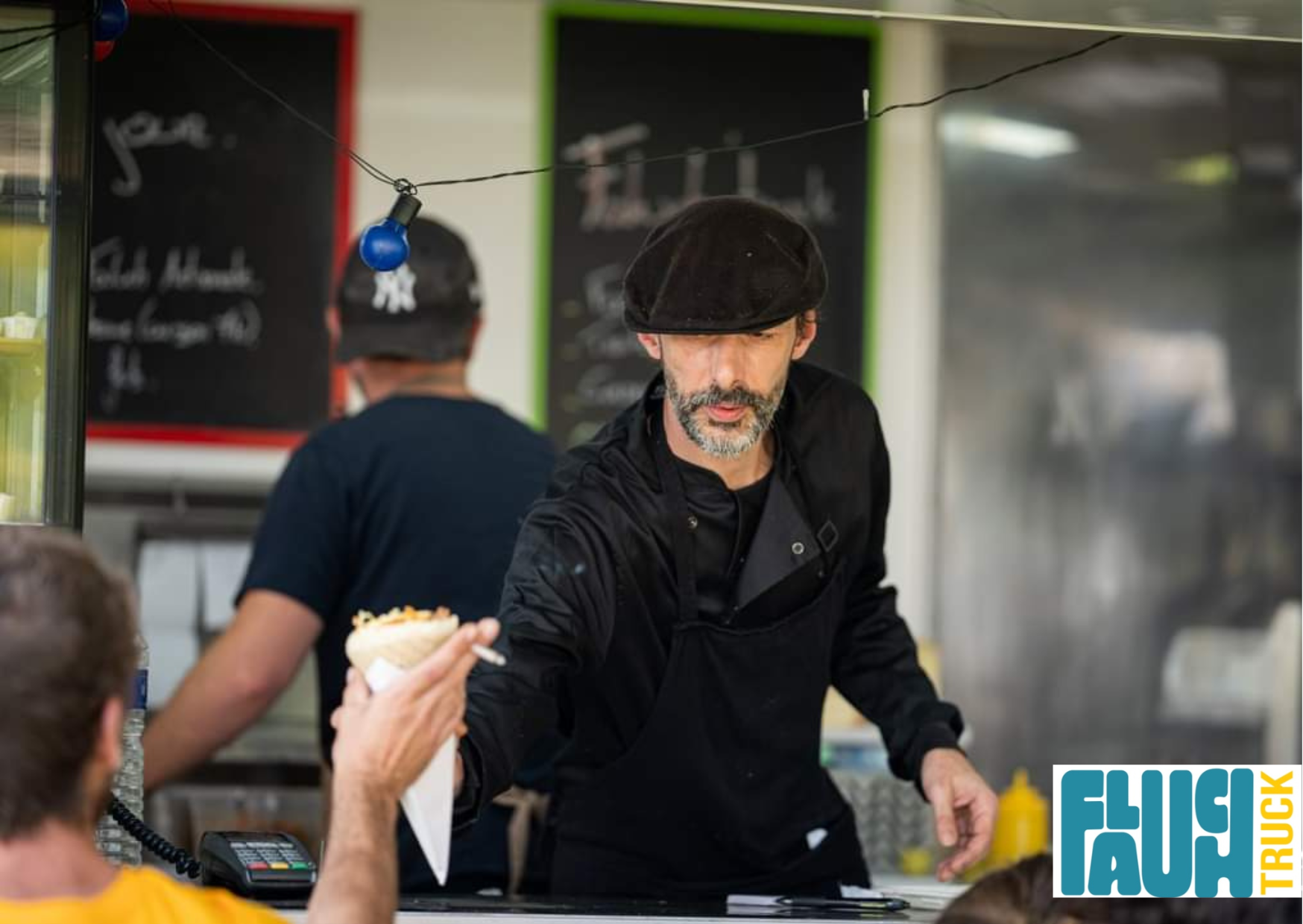 Un homme avec une barbe et un chapeau noir, probablement un chef ou vendeur, sert un sandwich à un client dans un restaurant ou un stand de nourriture. Il y a un tableau noir en arrière-plan avec des écrits en français et un éclairage suspendu avec u