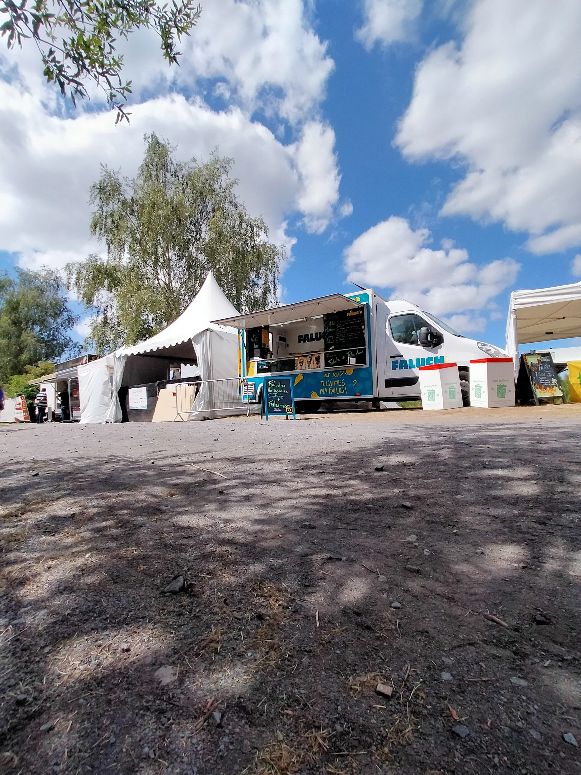 Un camion food truck bleu et blanc avec la marque "Faluch'Truck" stationné dans un marché en plein air, sous un ciel bleu parsemé de nuages, entouré de stand de vente avec des enseignes