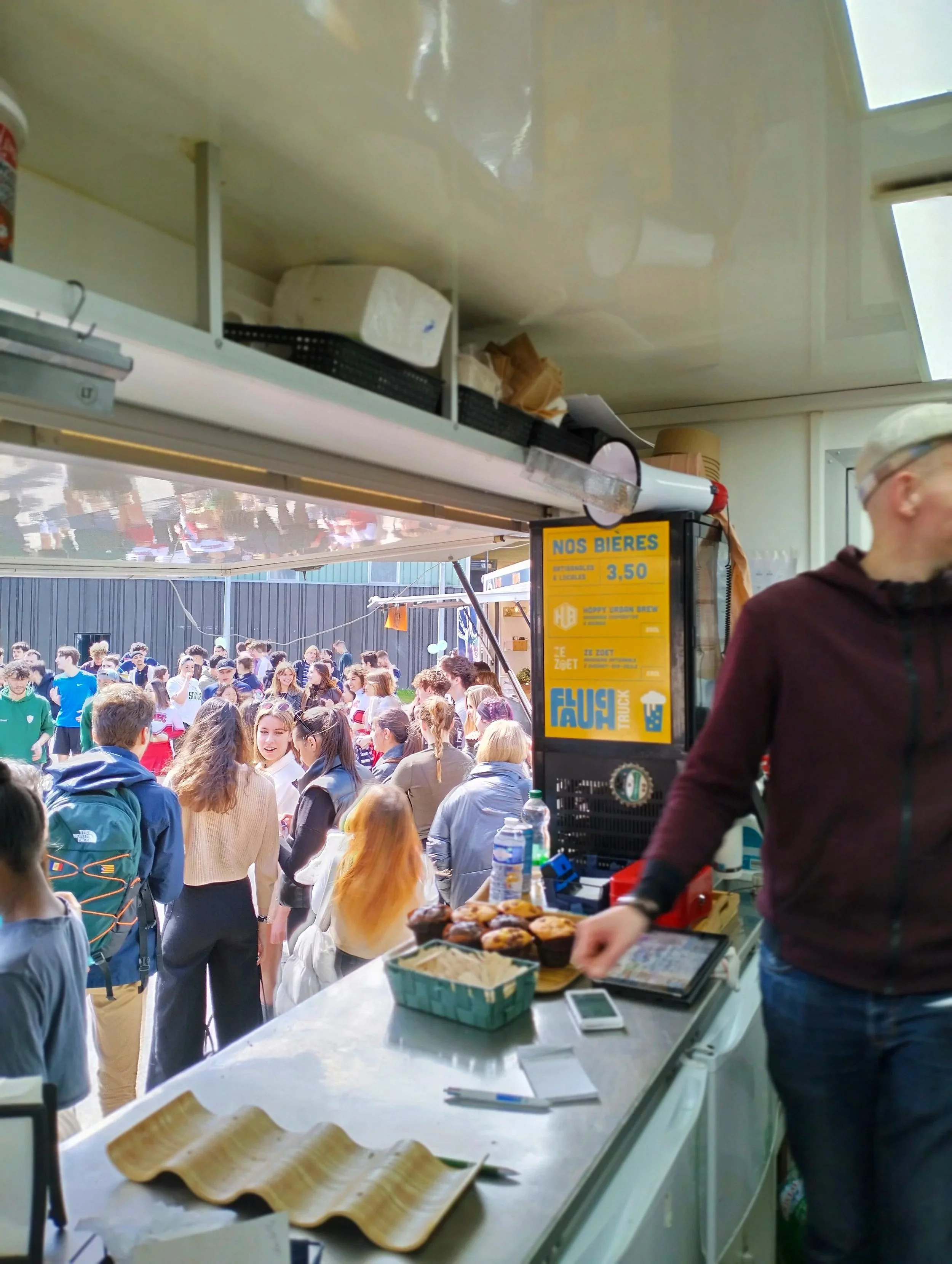 Dans le faluch'truck lors d'un événement, avec beaucoup de personnes à l'extérieur, certains attendant ou discutant, et des muffins ou gâteaux sur le comptoir, avec un homme portant des lunettes et un bonnet à droite.