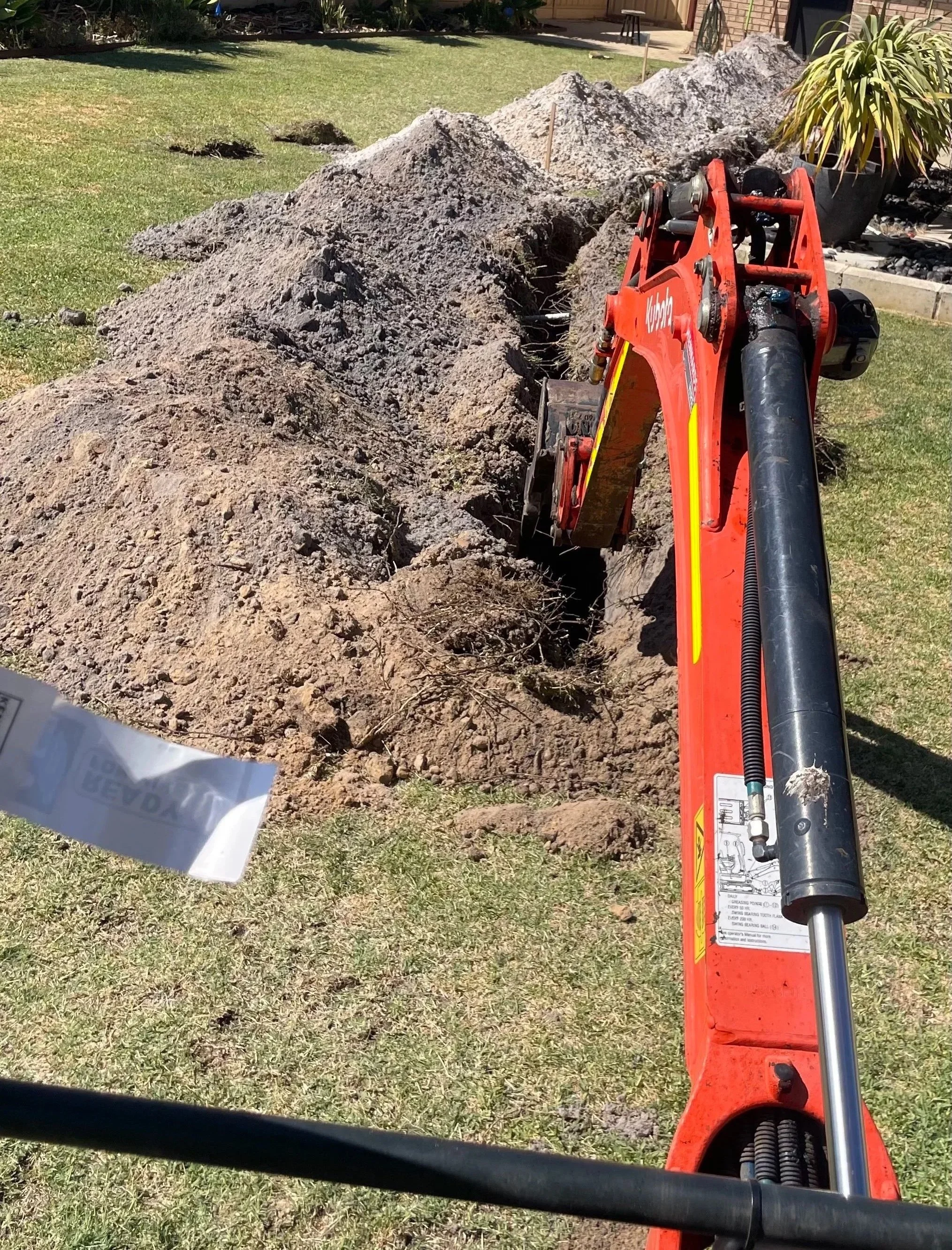Excavator digging a trench in a backyard with grass, plants, and a brick house in the background.