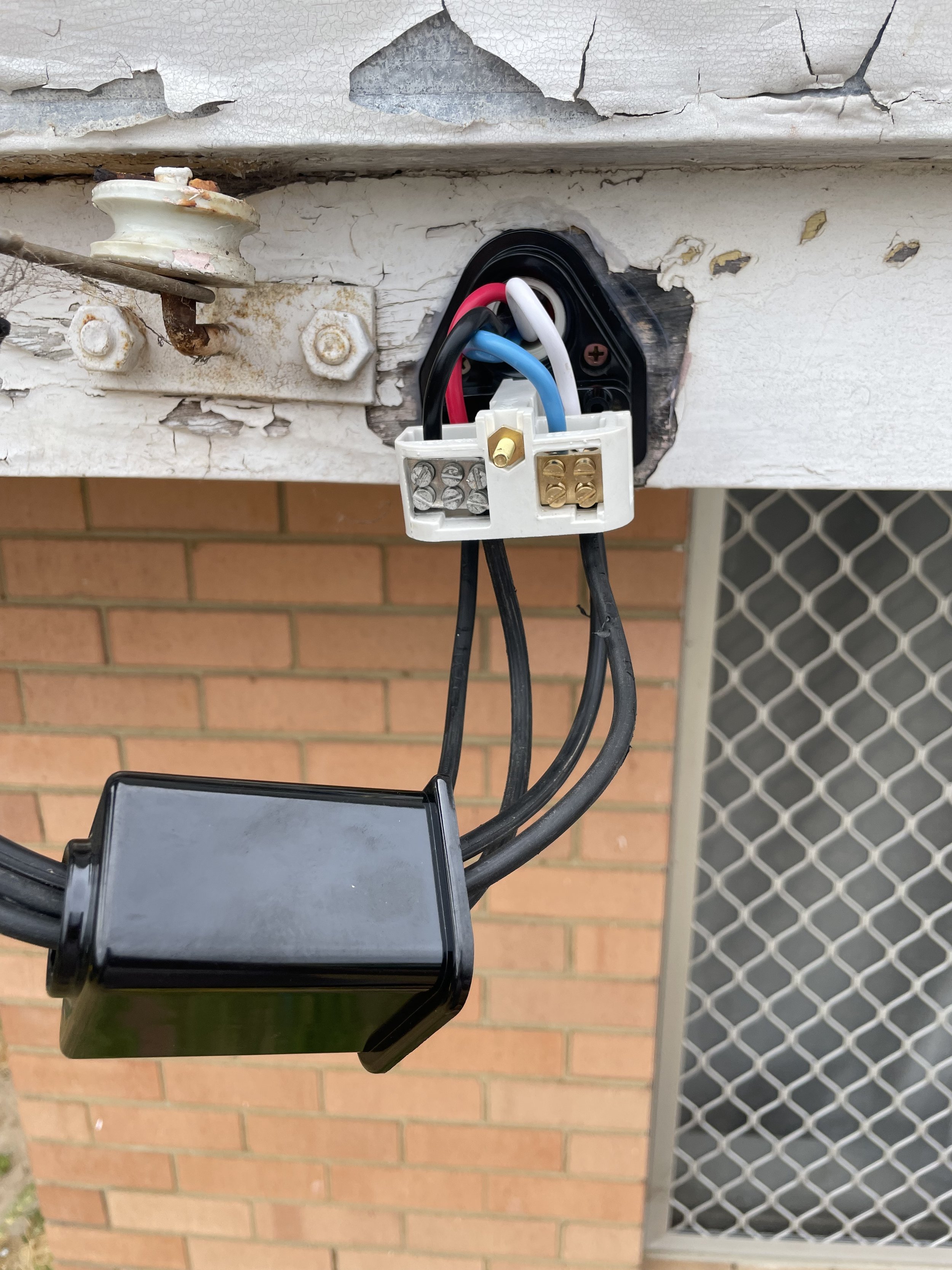 Close-up of an electrical outlet with wires connected, mounted on a cracked and peeling white wall, with a brick wall and metal gate in the background.
