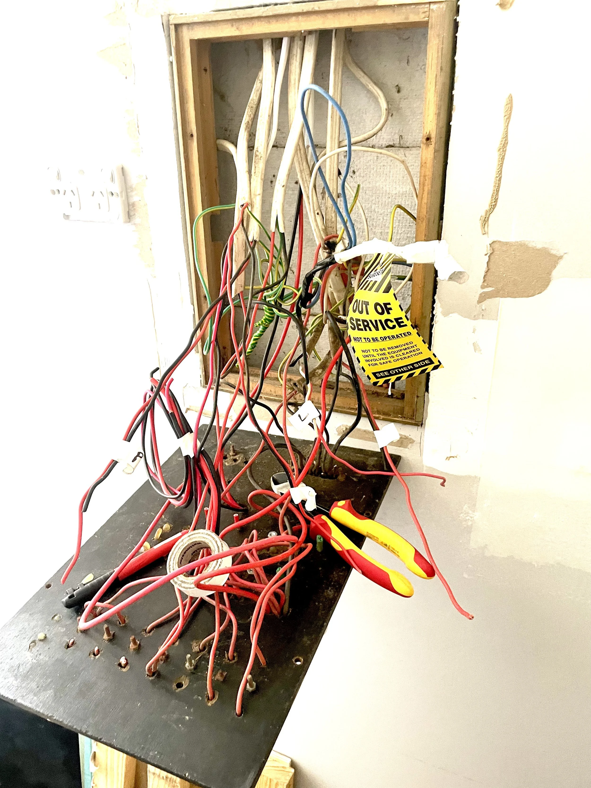 An open electrical panel with tangled wires, a pair of yellow and red-handled pliers, and a yellow "Out of Service" tag hanging on the wires.