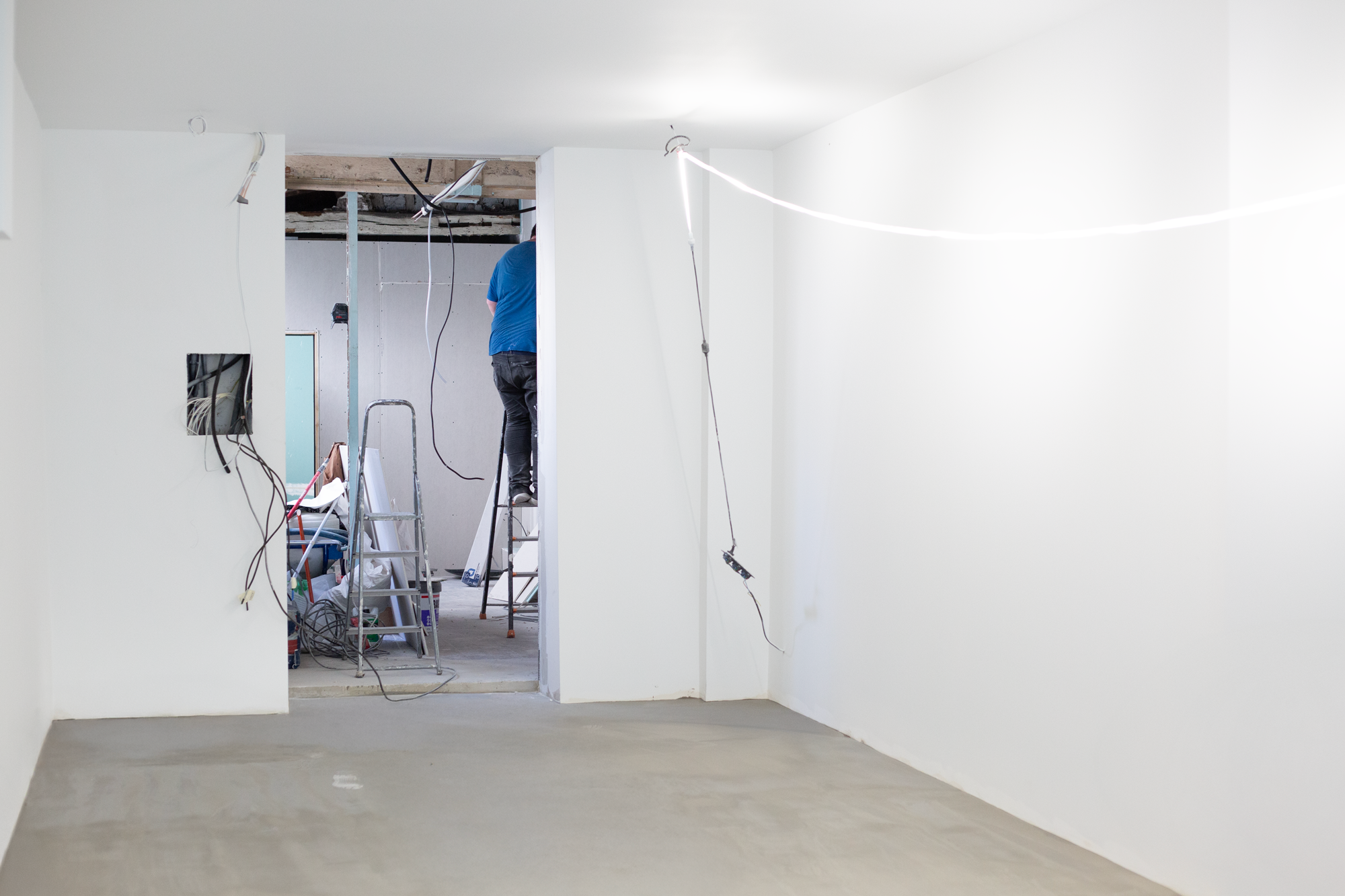 Une pièce en cours de rénovation avec un mur blanc, un sol en béton fraîchement coulé, et un espace de passage menant à une autre pièce où un homme travaille sur un échafaudage.