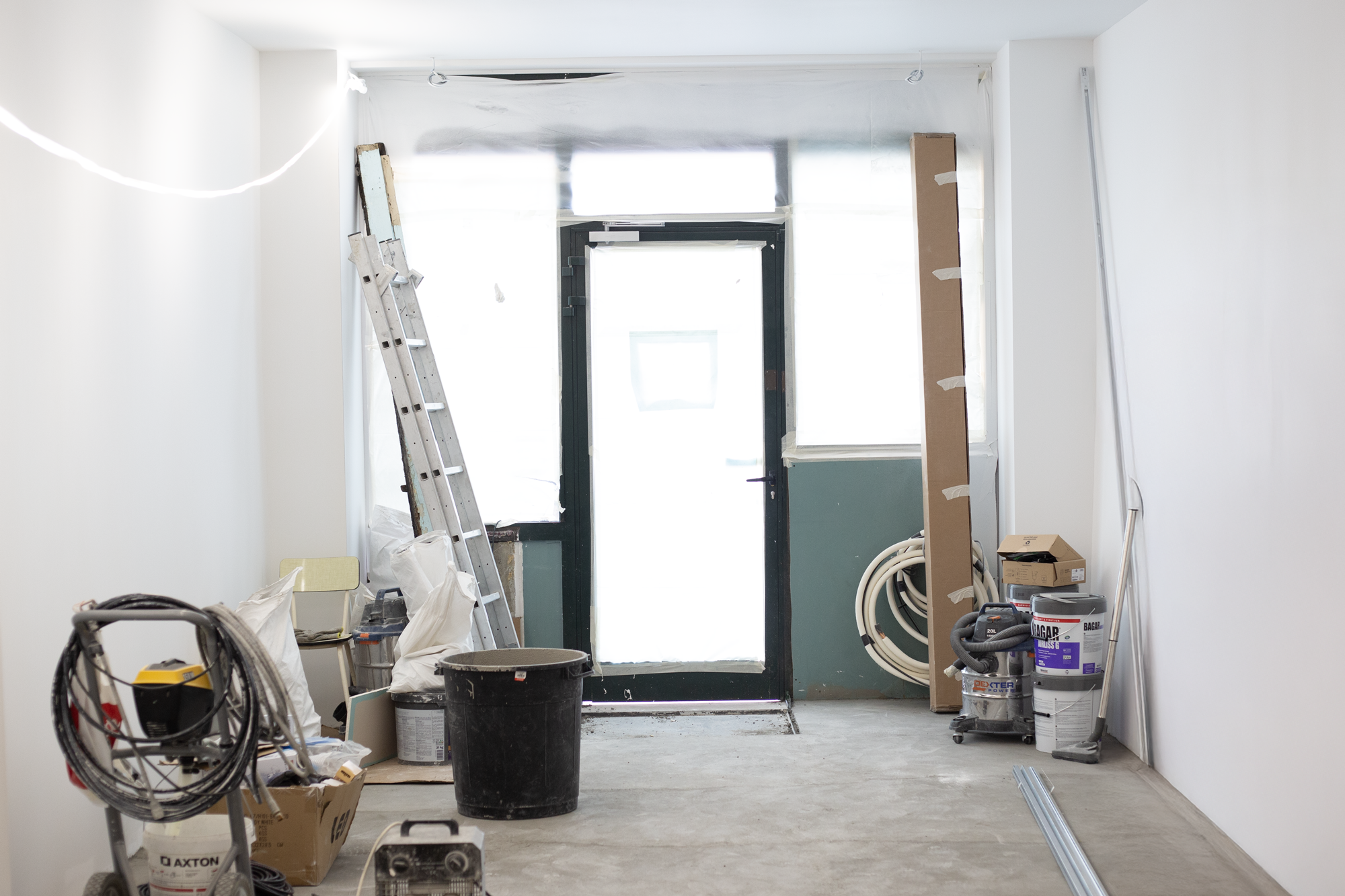 Une pièce en cours de rénovation avec des outils, des matériaux, un escabeau, une poubelle, des tuyaux et des peintures dans une salle blanche.