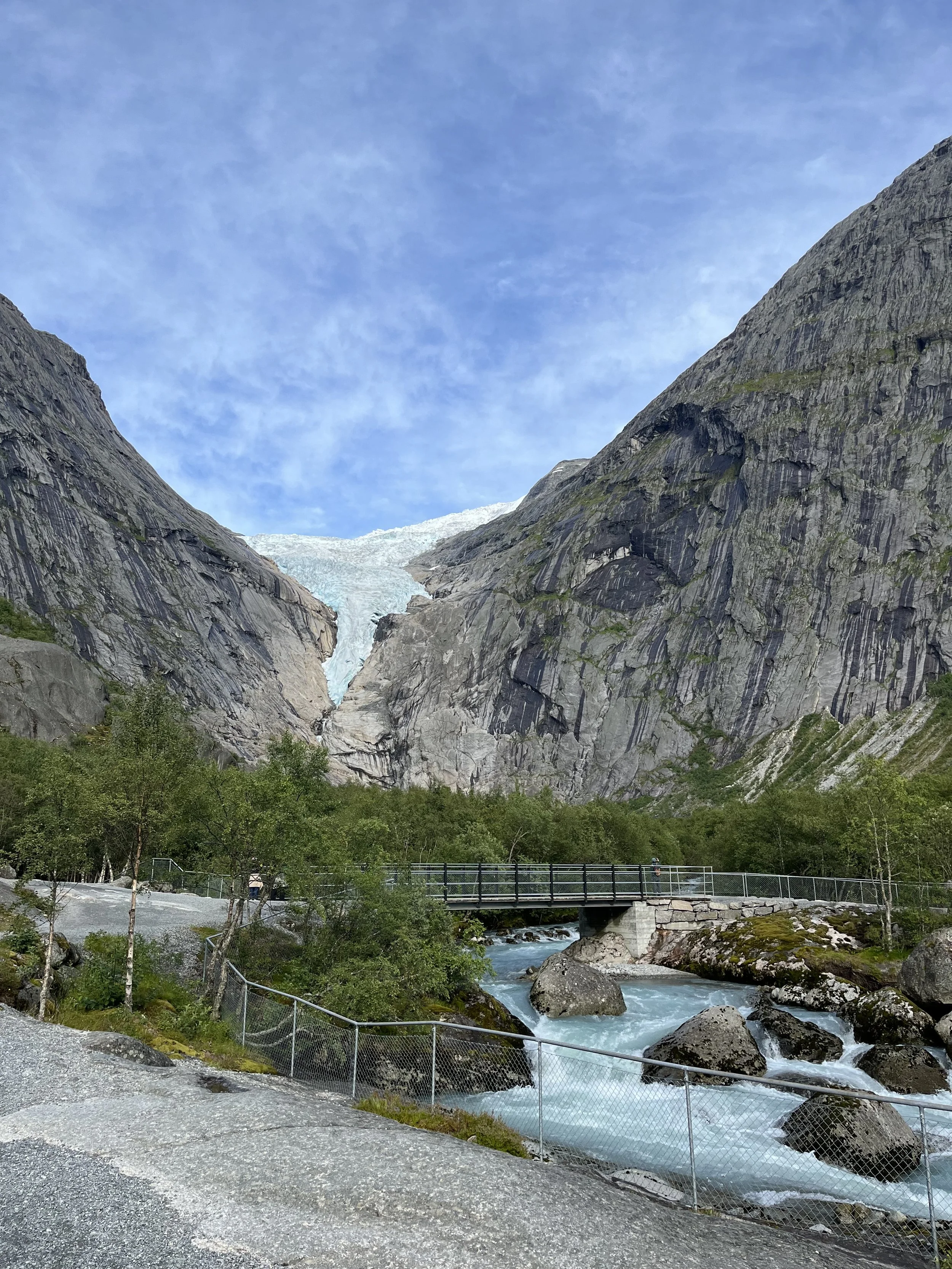 Scenic view of a mountain valley with a glacier nestled between steep rocky cliffs, a small bridge over rushing water, and lush green trees in the foreground under a partly cloudy blue sky.