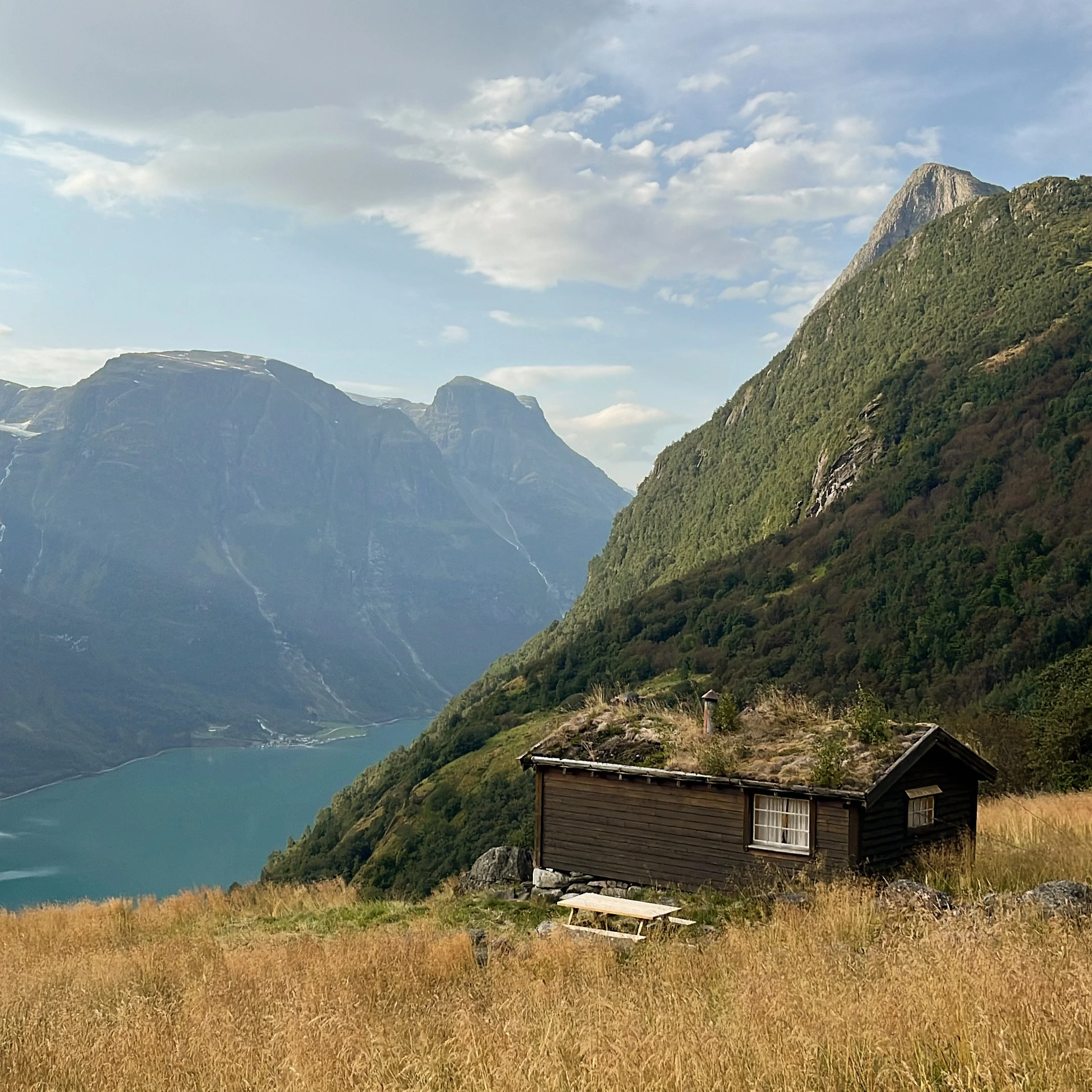 A small wooden cabin with a grass roof sits on a grassy hillside overlooking a fjord, surrounded by green mountains under a partly cloudy sky.