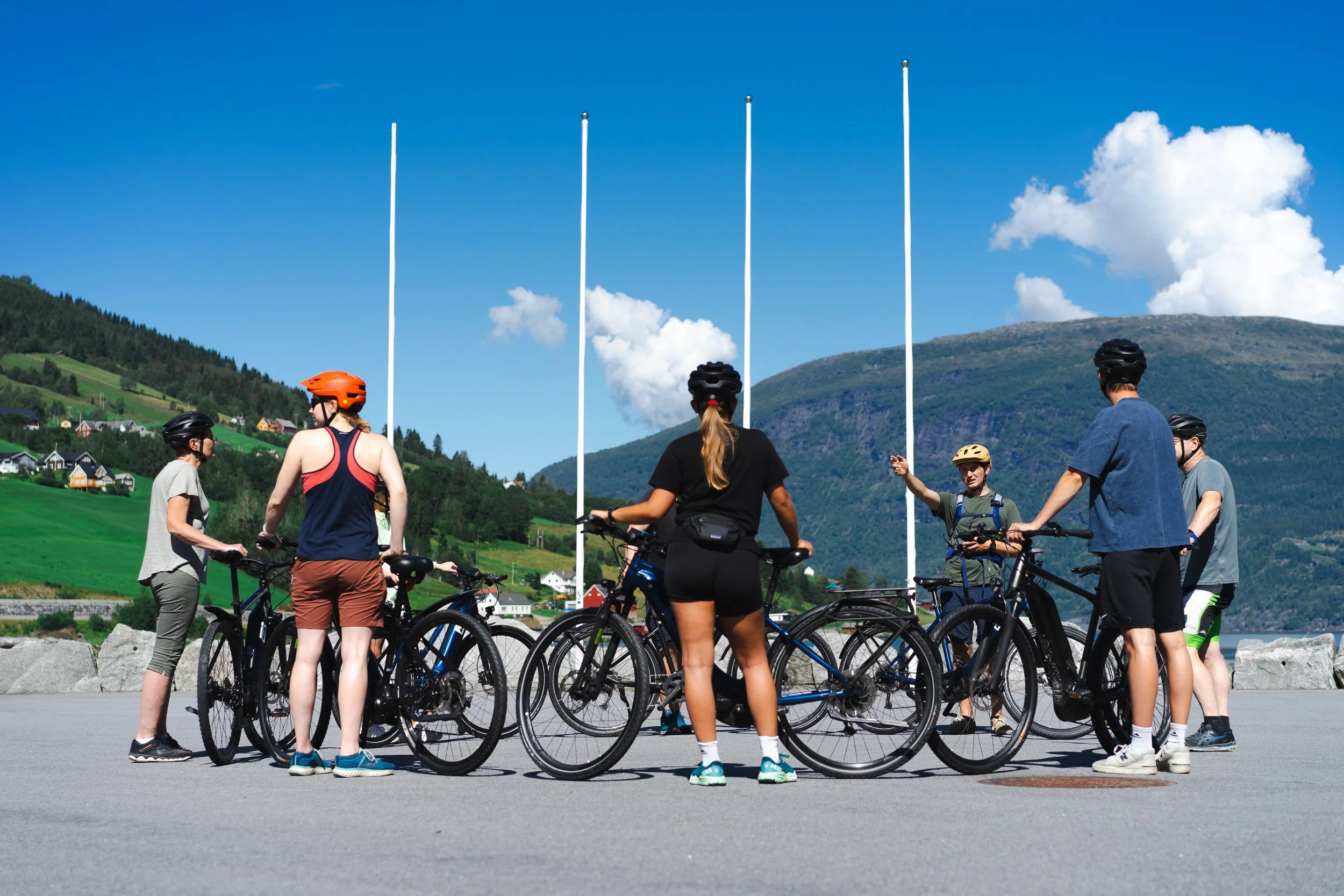 A group of eight cyclists gathered in a circle with their bikes outdoors on a sunny day. They are wearing helmets and casual athletic clothing, with a scenic background of mountains, green hills, and a blue sky with clouds.
