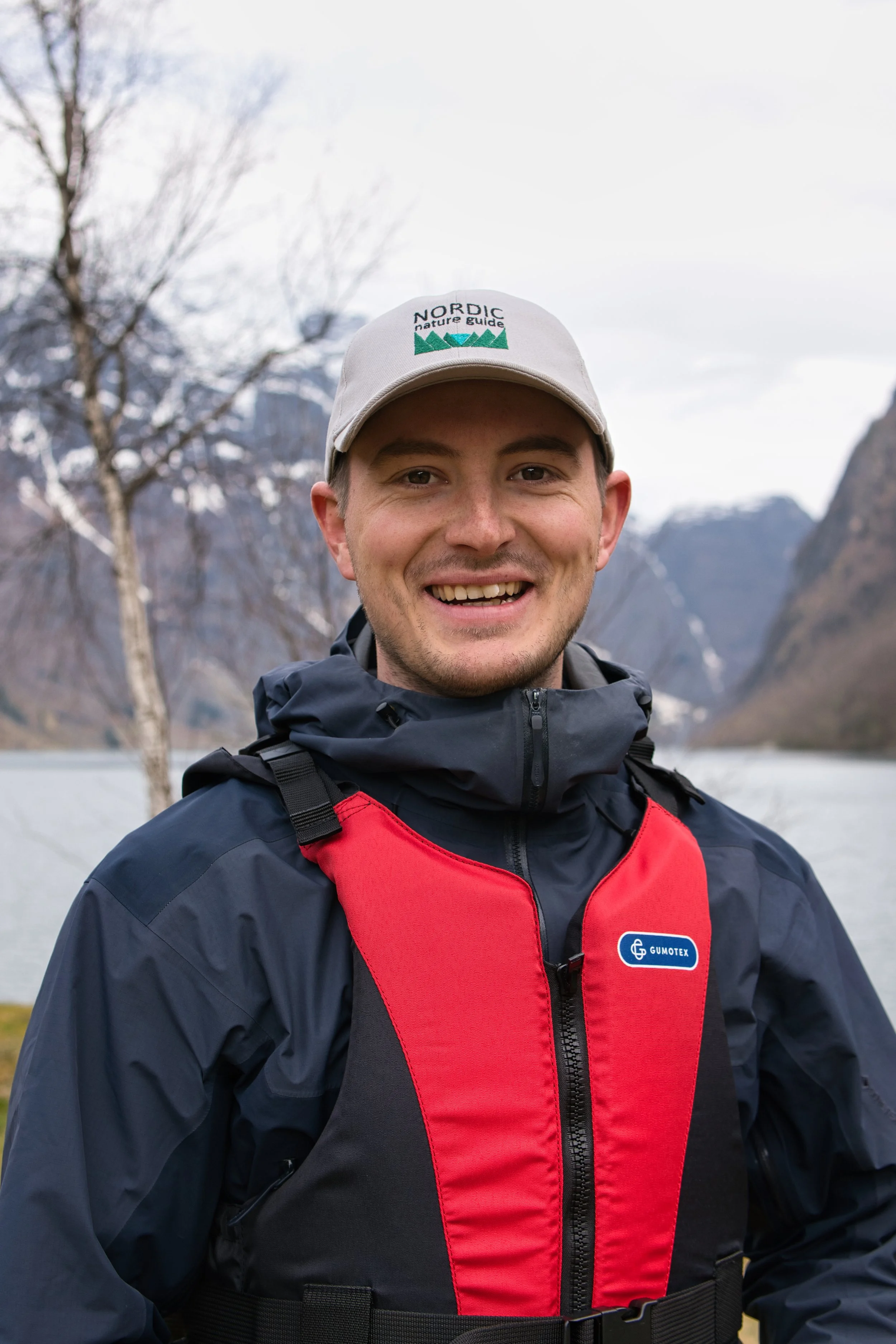 A smiling young man wearing a gray cap with green and black text and logo, a black and red jacket with a blue logo, outdoors with mountains, a leafless tree, and a river in the background.