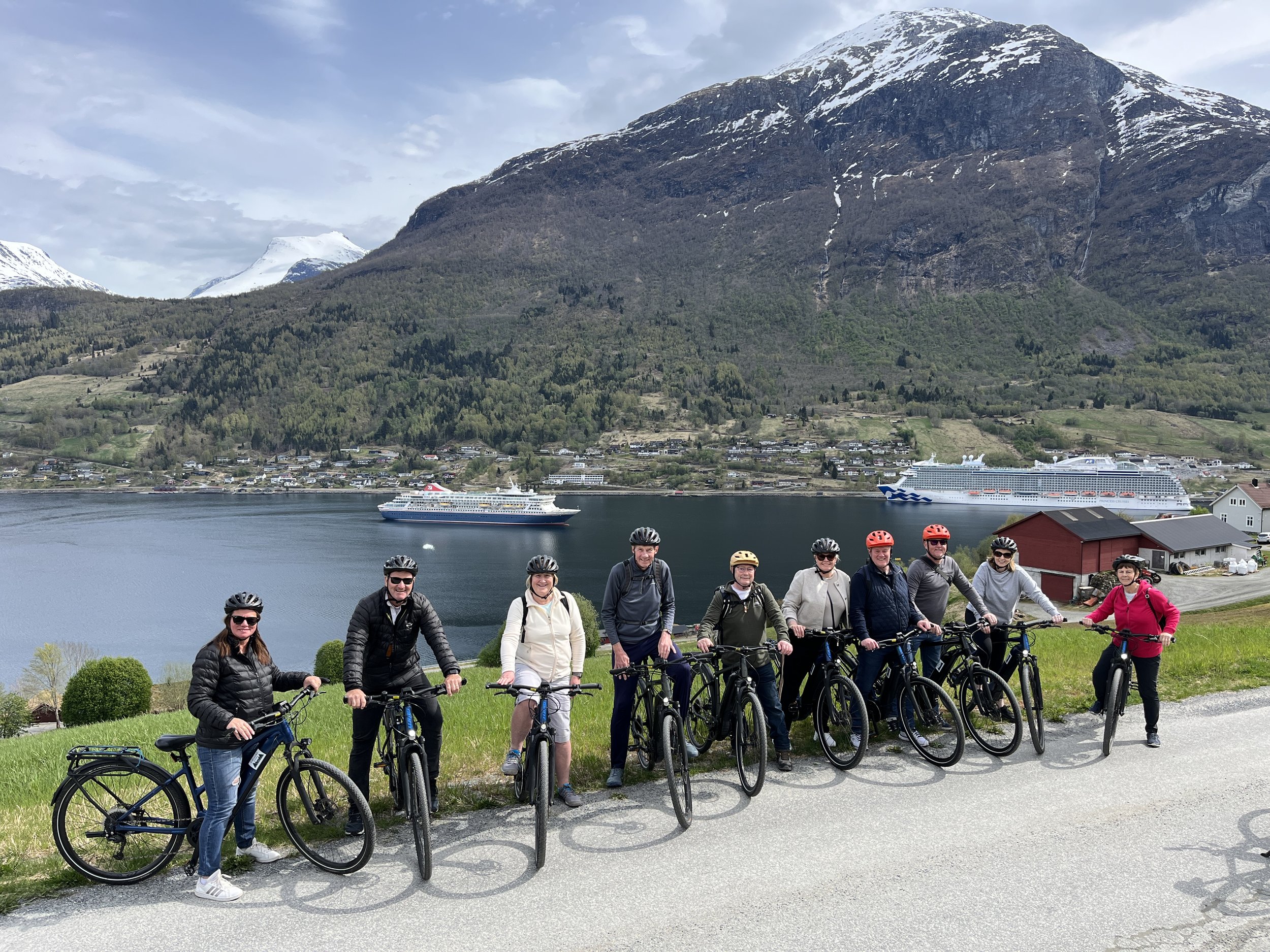 A group of ten people wearing helmets with bicycles, standing on a scenic road with a lake, mountains, and cruise ships in the background.