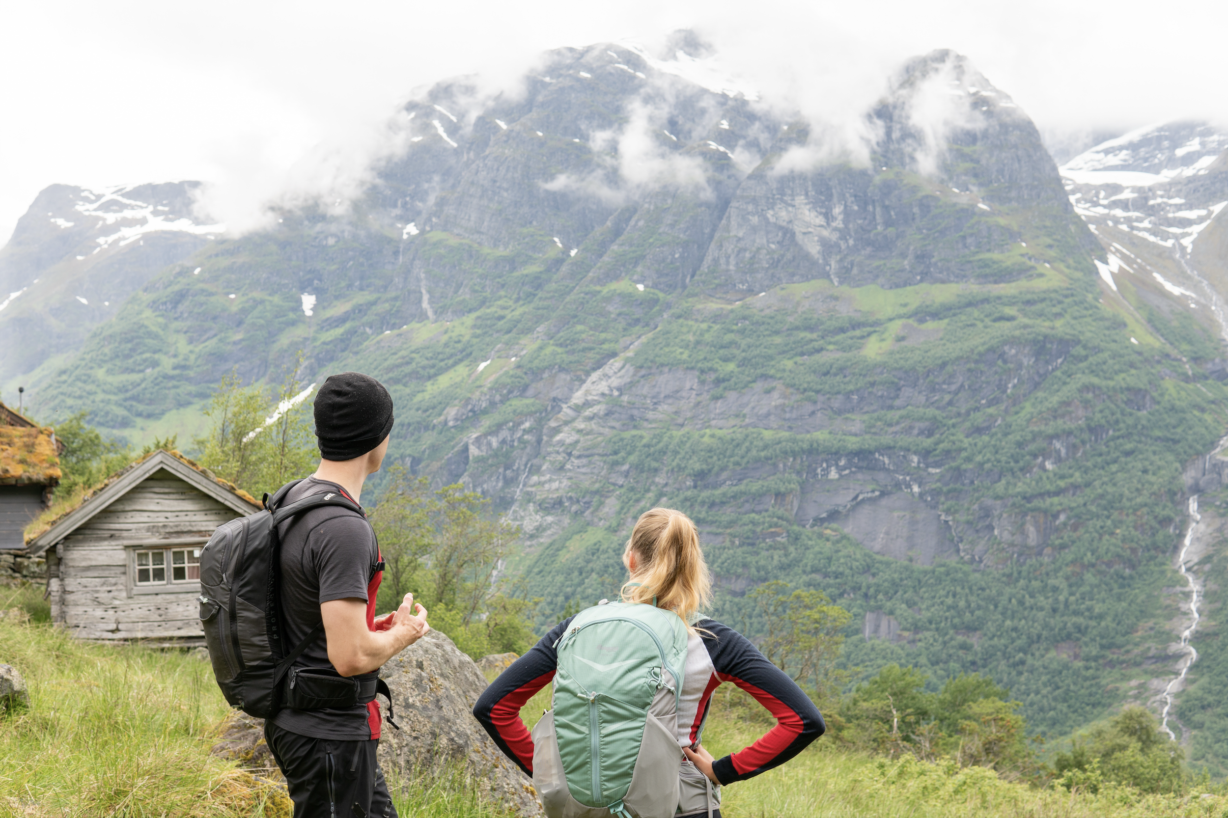 Two hikers, a man and a woman, stand on a grassy field looking at mountains with green slopes, rocky faces, and snow patches, with cloudy weather.