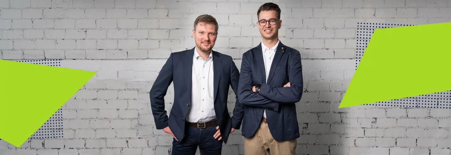 Zwei junge Männer in Anzügen stehen vor einer weißen Ziegelwand, lächeln und schauen in die Kamera.