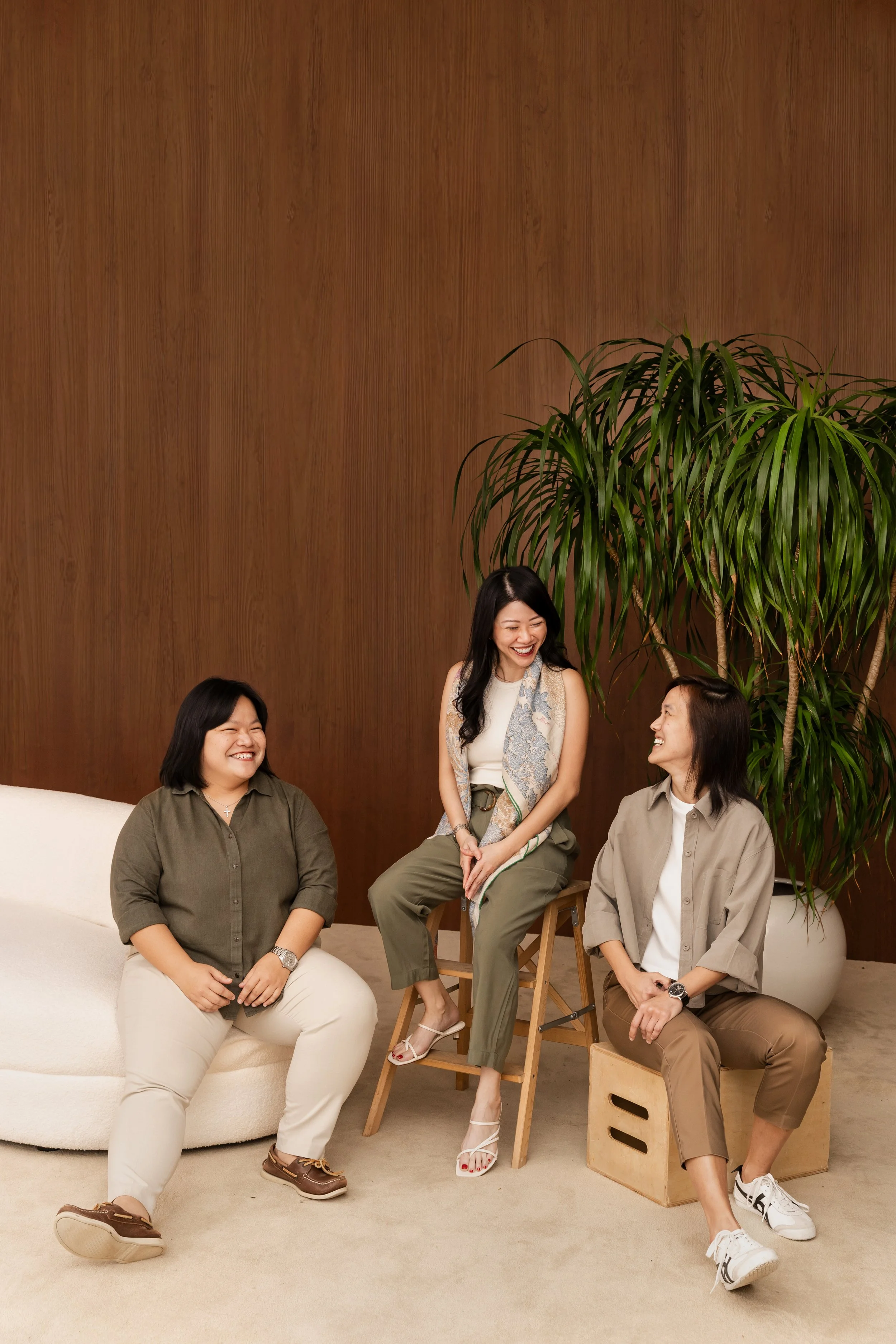 A group of five women sitting and standing in a casual, indoor setting with a wooden wall and a large potted plant behind them, all smiling and engaging in conversation.