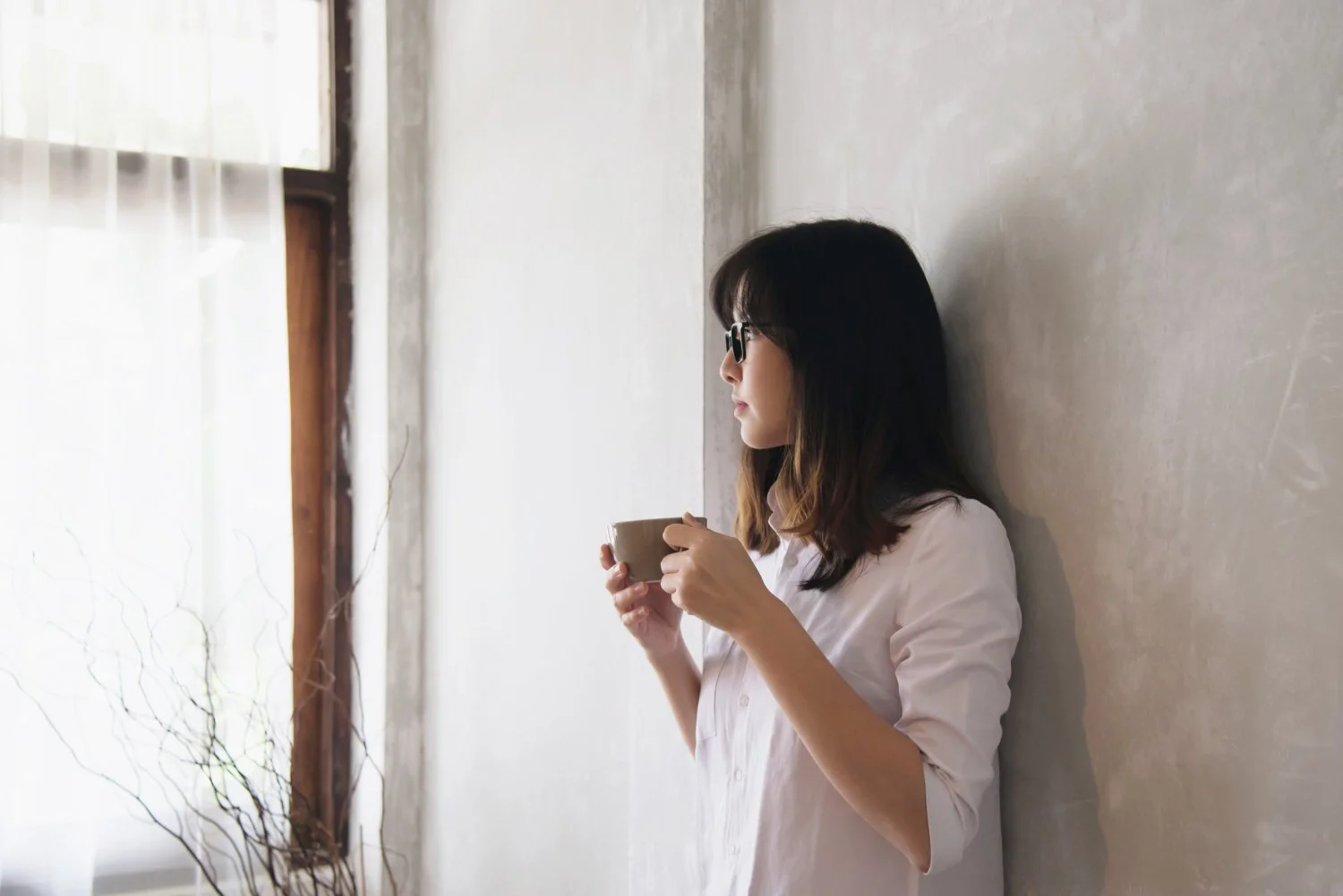 A woman with dark, shoulder-length hair and sunglasses, wearing a white shirt, stands against a plain wall, holding a mug and gazing out of a window.