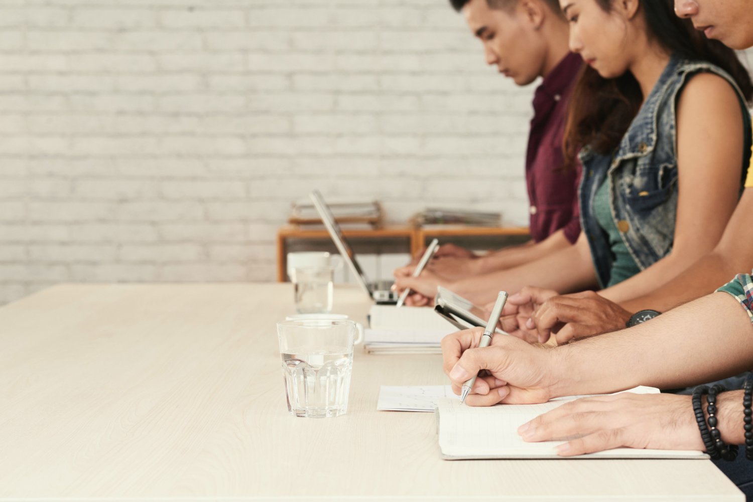People sitting at a conference table taking notes with a glass of water in front of them, in a professional setting.