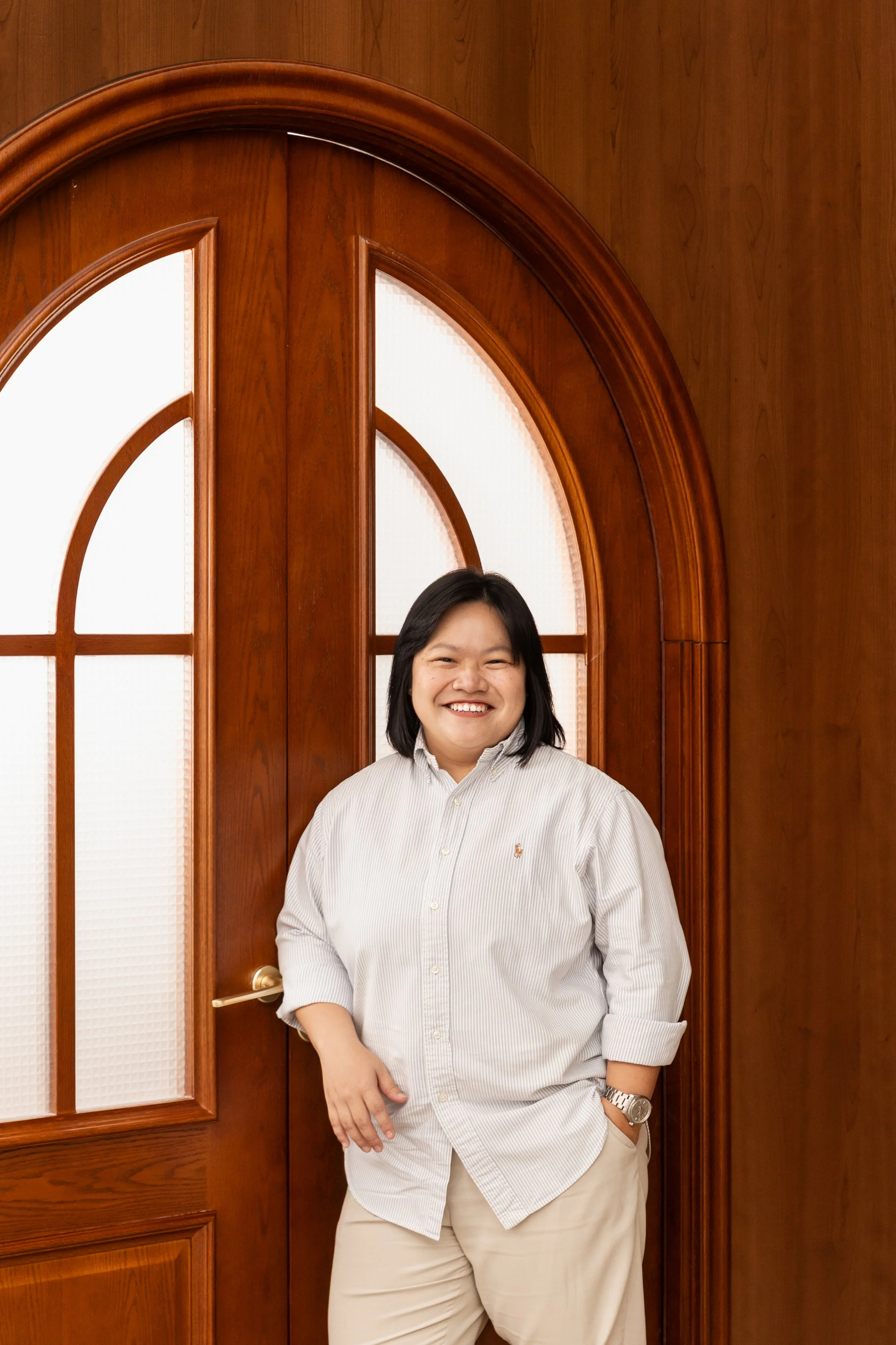 A person with shoulder-length black hair, wearing a white striped button-down shirt and beige pants, smiling and standing in front of a wooden door with glass panels.