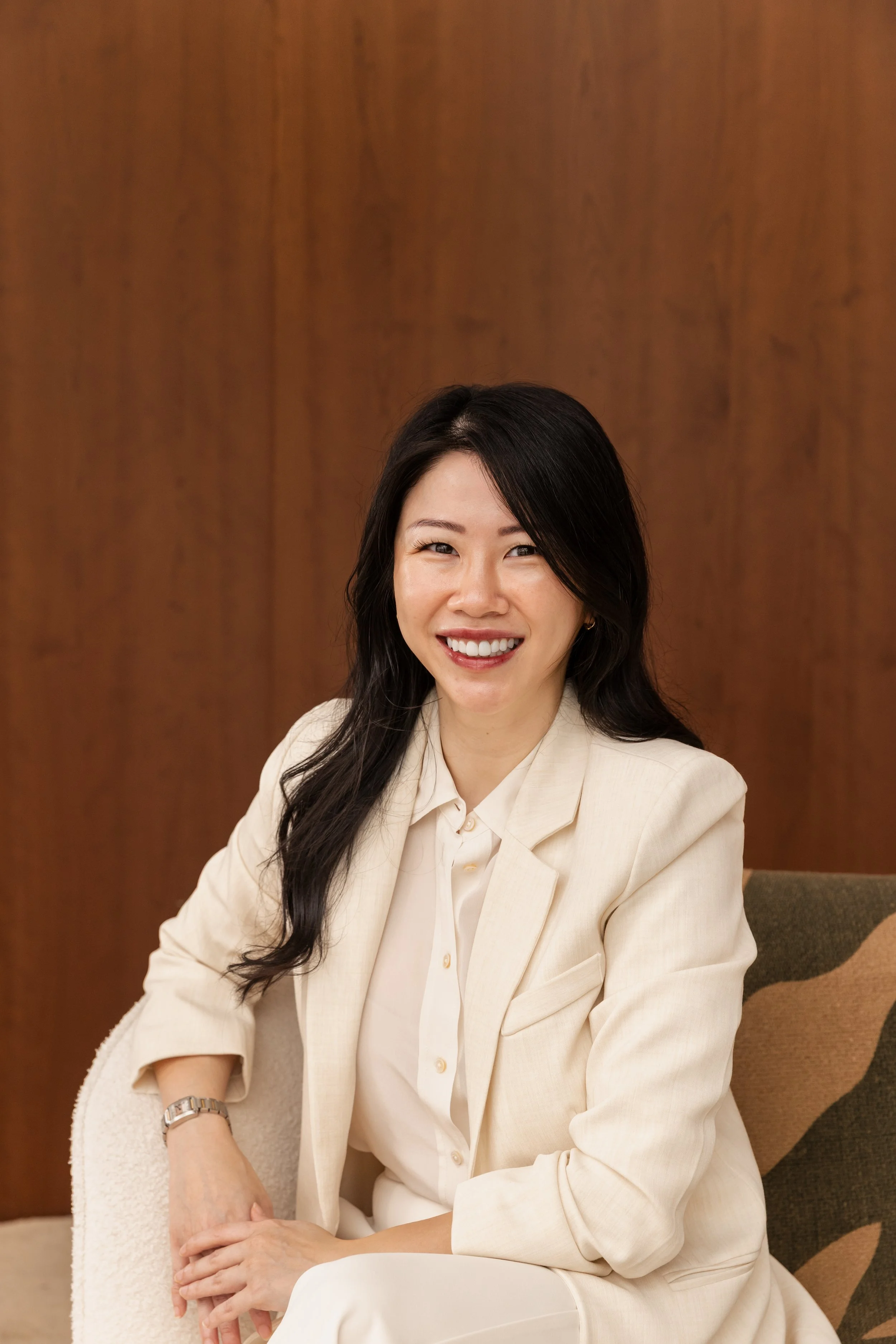 A woman with long black hair smiling, wearing a cream-colored blazer over a white shirt, sitting on a brown chair against a wood-paneled wall.