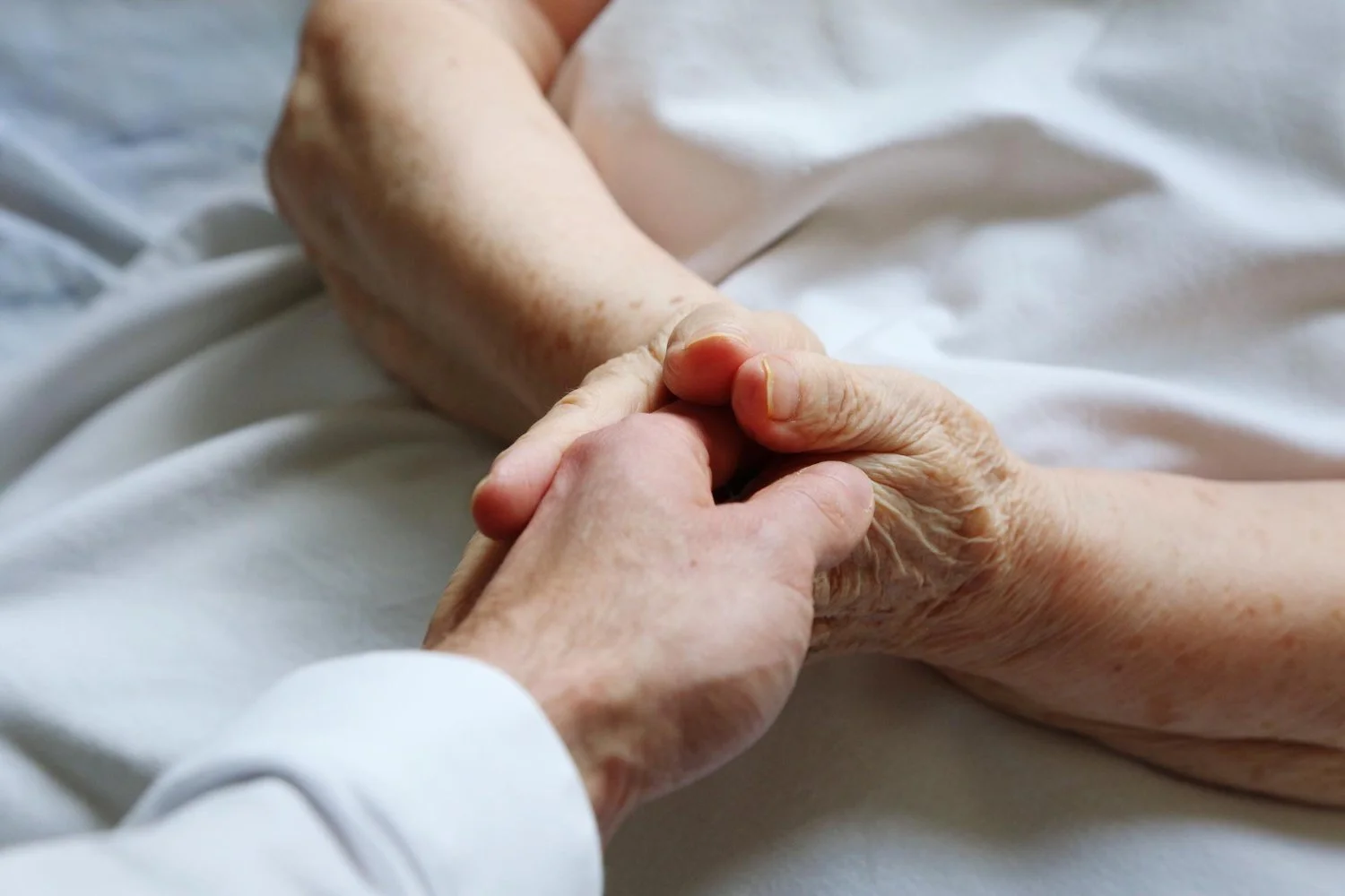 A close-up of two hands holding each other, one appears to be a young adult's and the other an elderly person's, on a bed with white sheets.