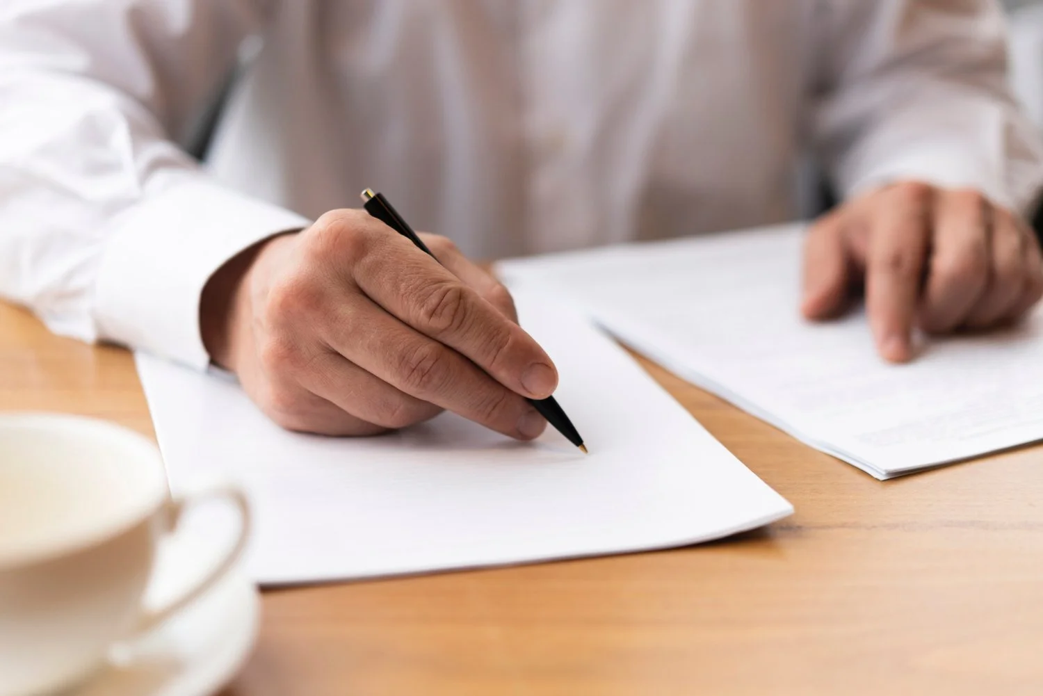 A person in a white dress shirt is writing on a sheet of white paper with a black pen, with additional papers on a wooden desk and a white coffee mug nearby.