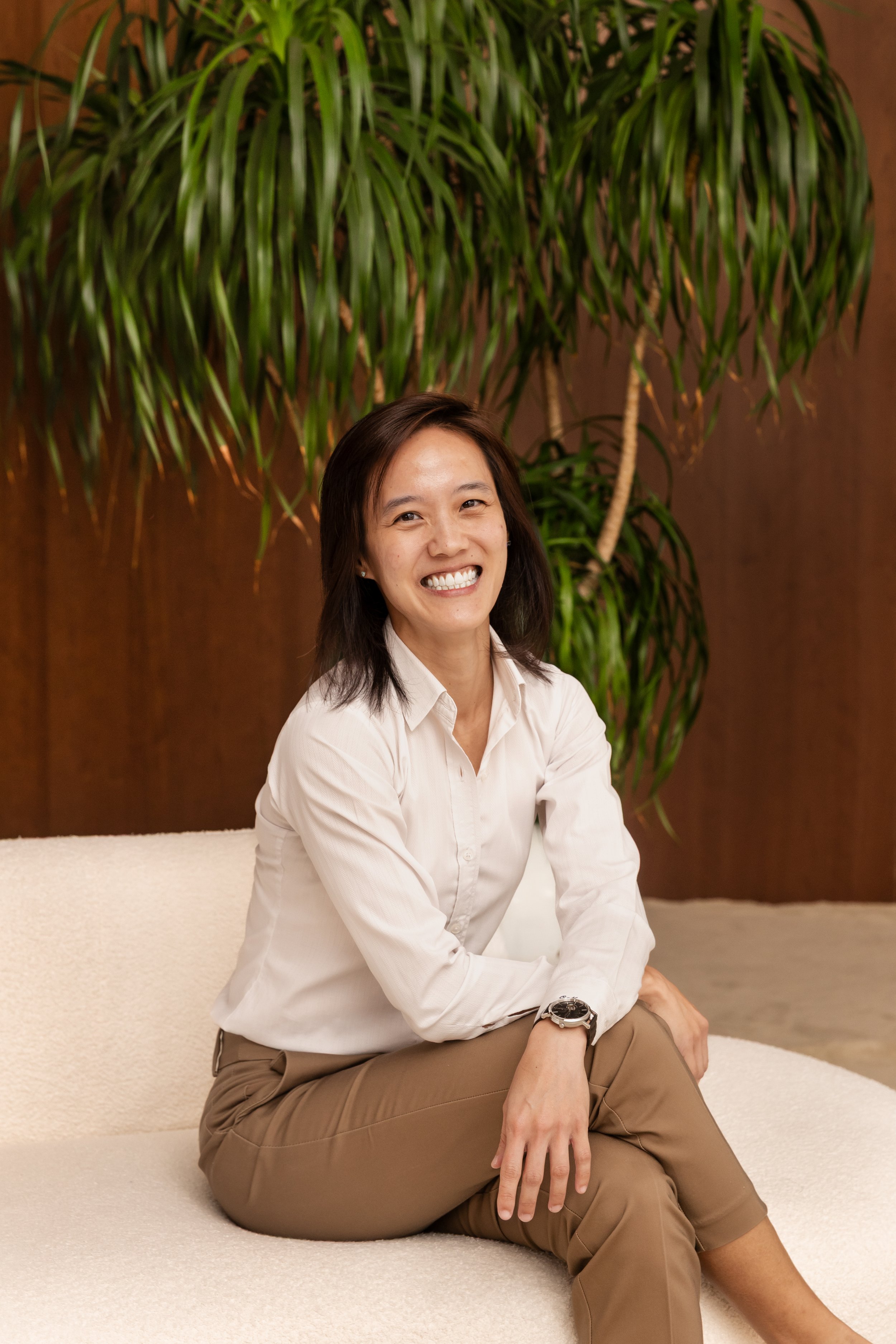 A woman sitting on a beige sofa with a large green plant behind her, smiling broadly with teeth showing, wearing a white shirt, tan pants, and a watch on her left wrist.