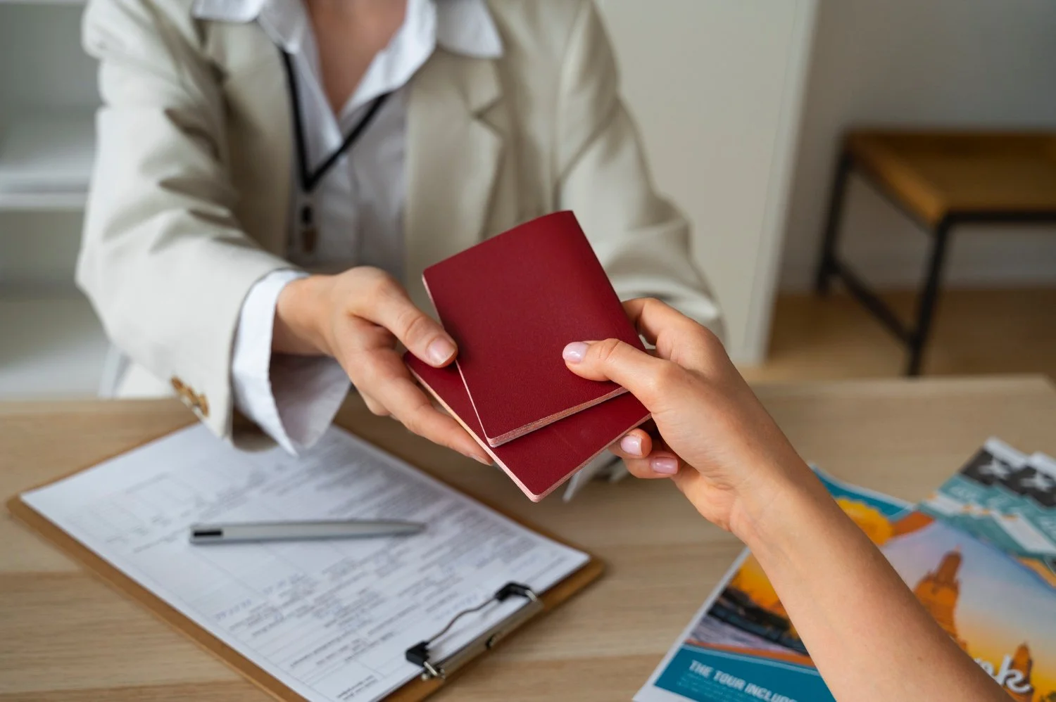 A person in a medical coat hands a red passport to another person at a desk with a clipboard, a pen, and travel brochures.
