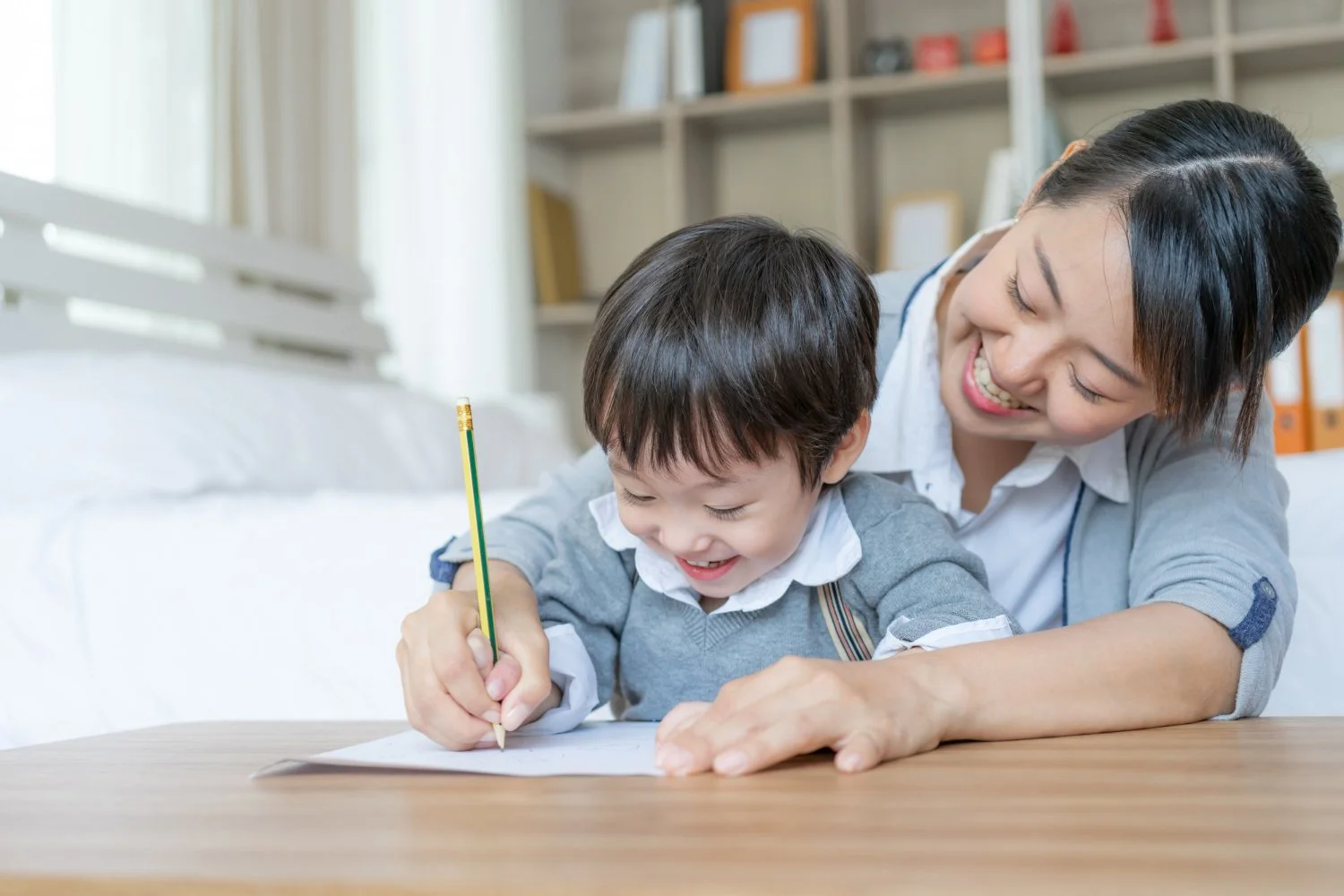 A smiling woman helping a young boy with a drawing or writing activity at a wooden table in a bright room.