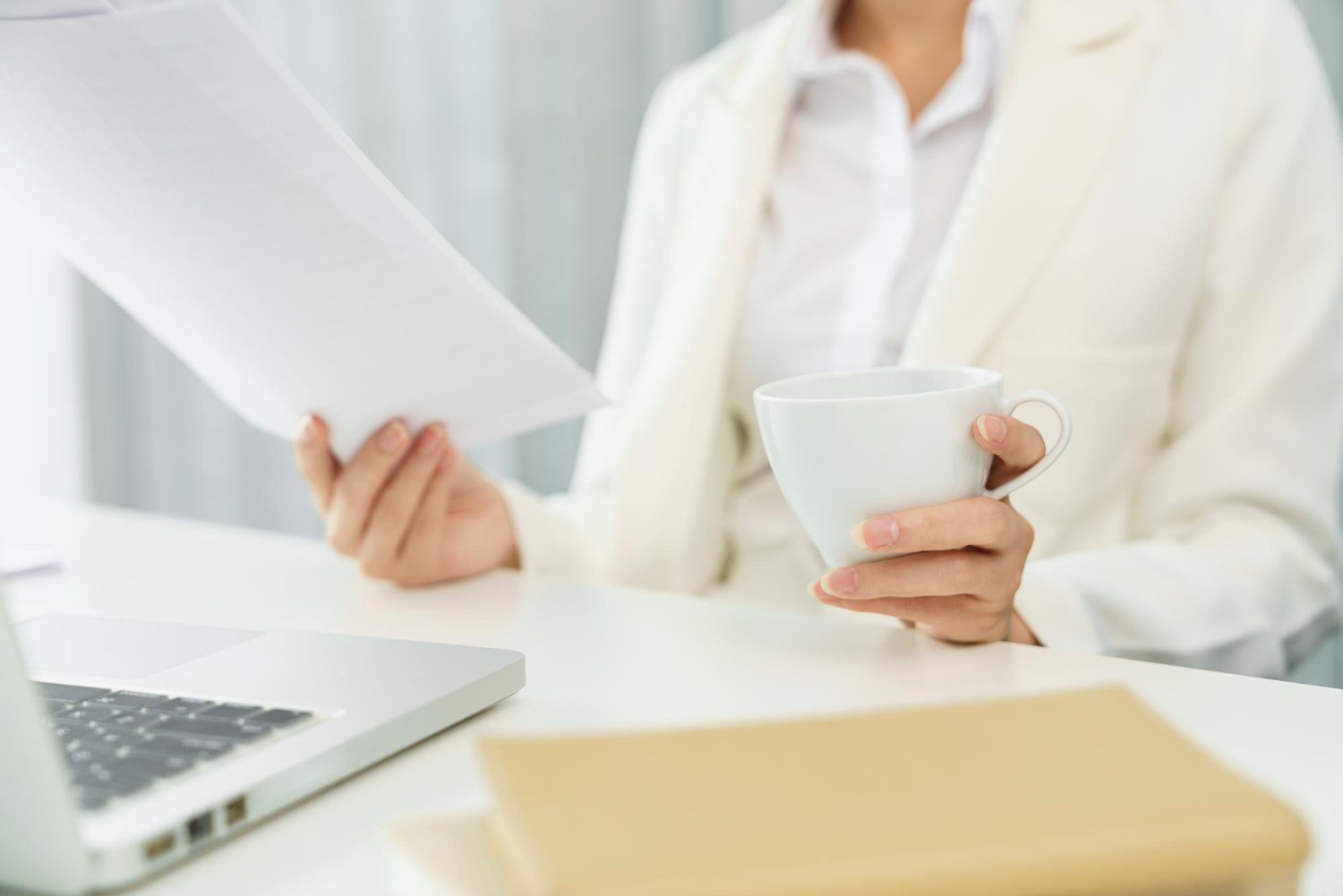 Person in white attire holding a white coffee cup in one hand and a paper in the other, sitting at a desk with a laptop and a yellow notebook.