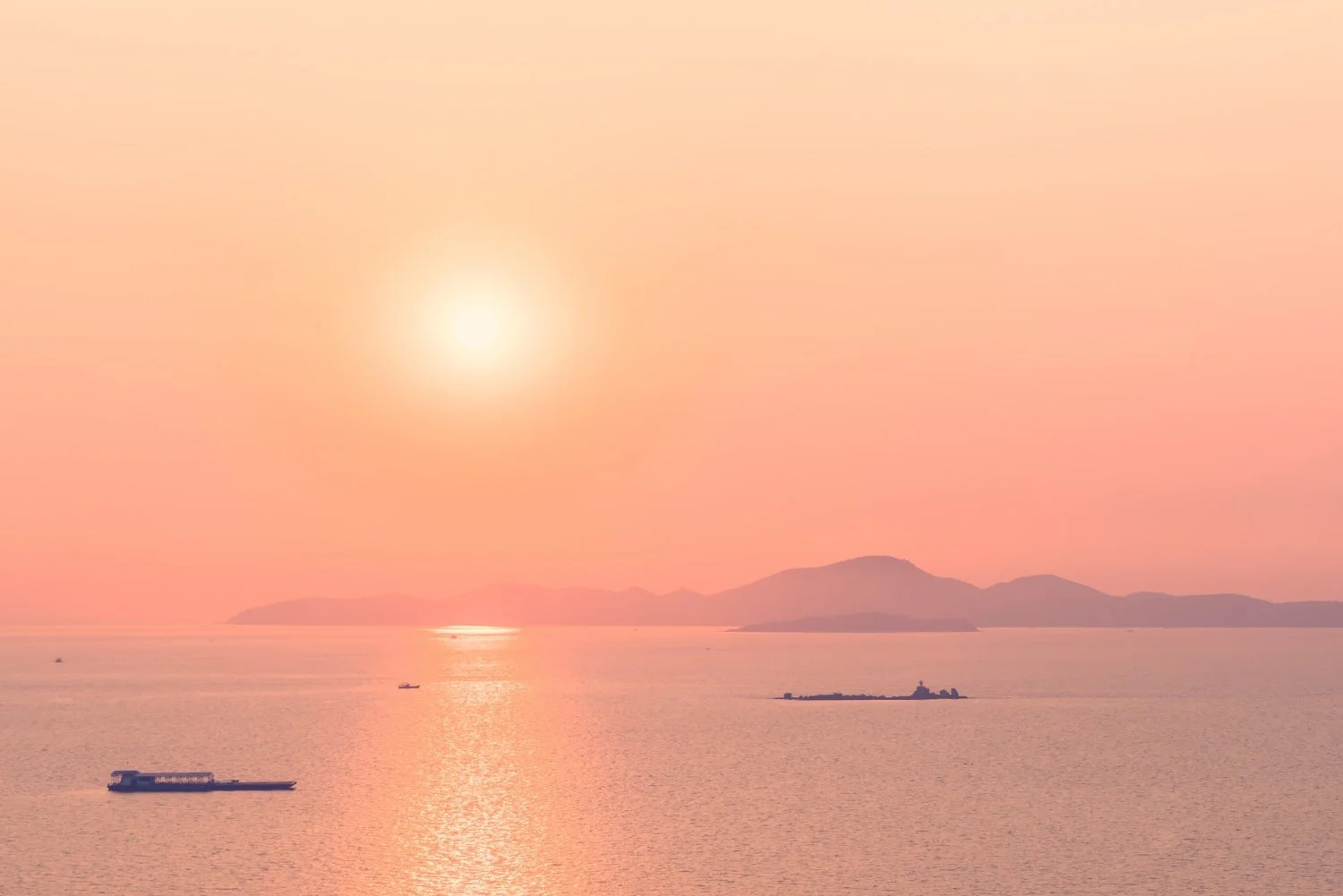 Sunset over calm ocean with mountains in the distance, small ships on the water, pinkish sky.