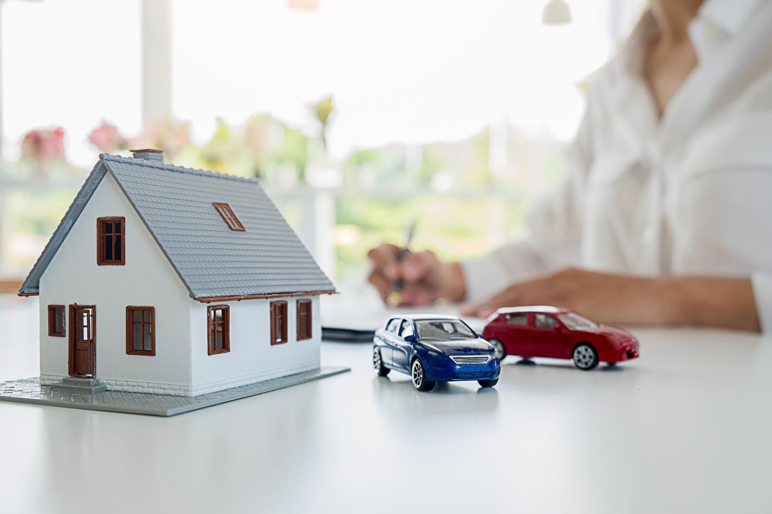 Model house with two toy cars on a table, with a person writing in the background.
