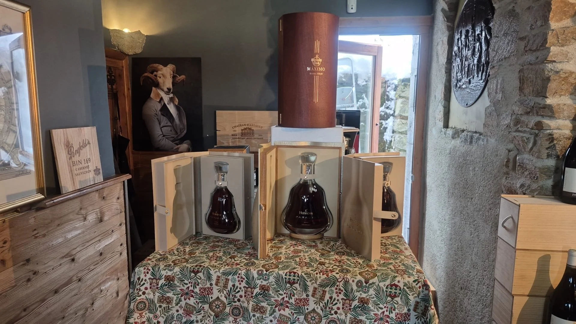 Décoration intérieure avec bouteilles de spiritueux dans des étuis en bois sur une table recouverte d'une nappe à motifs floraux, murs en pierre et objets décoratifs