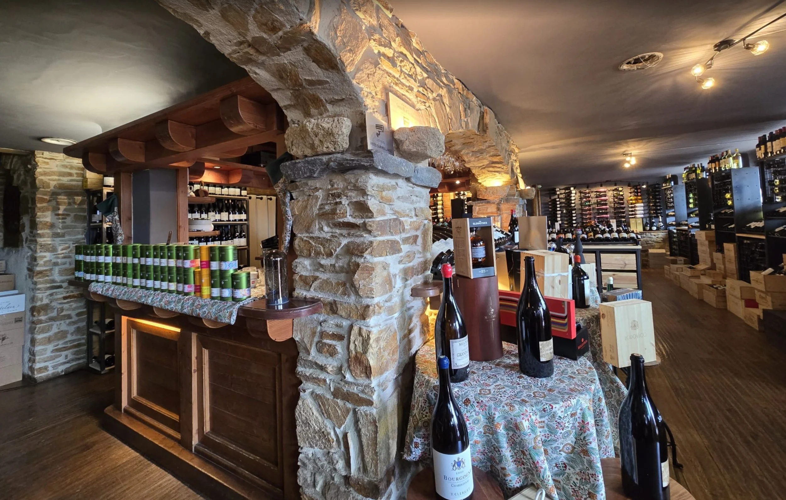 Intérieur d'une cave à vin avec étagères remplies de bouteilles, un comptoir en bois avec organisation de vins, et une table décorée de bouteilles et de caisses à vin en bois.