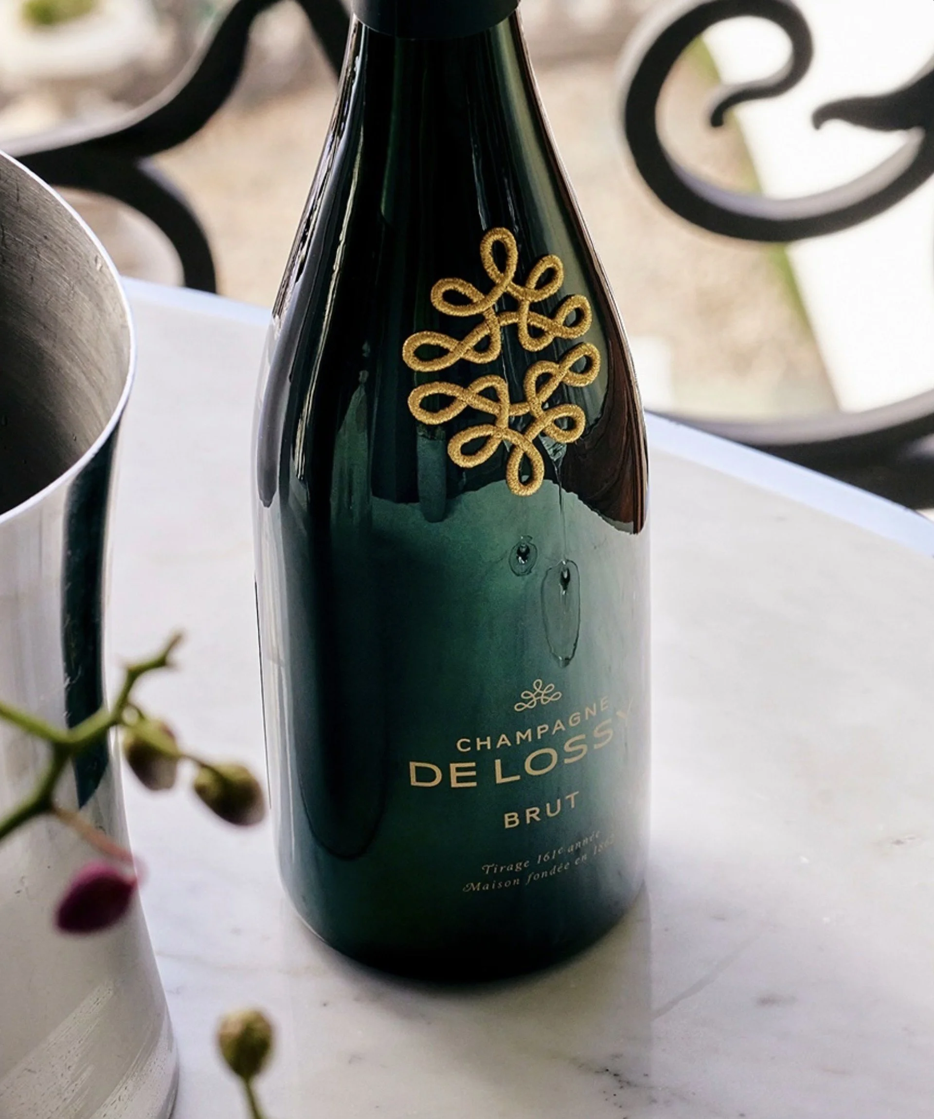 Bouteille de champagne brut "De Lossy" posée sur une table en marbre, avec un décor de chair de bouteille dorée en haut de la bouteille.