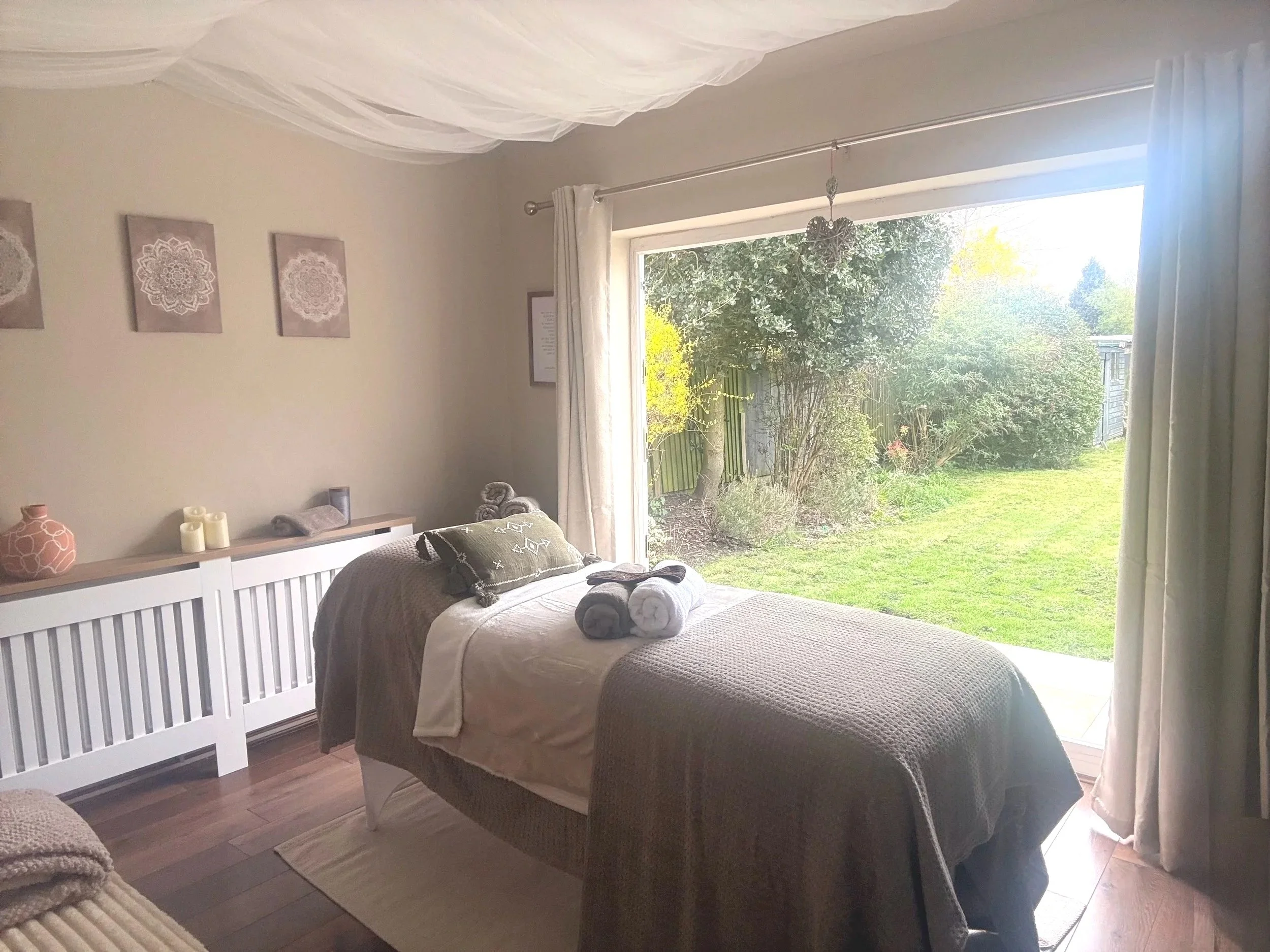 A cozy bedroom with a bed facing a large sliding glass door leading to a lush green garden. The room is decorated with neutral tones, wall art, and soft furnishings.