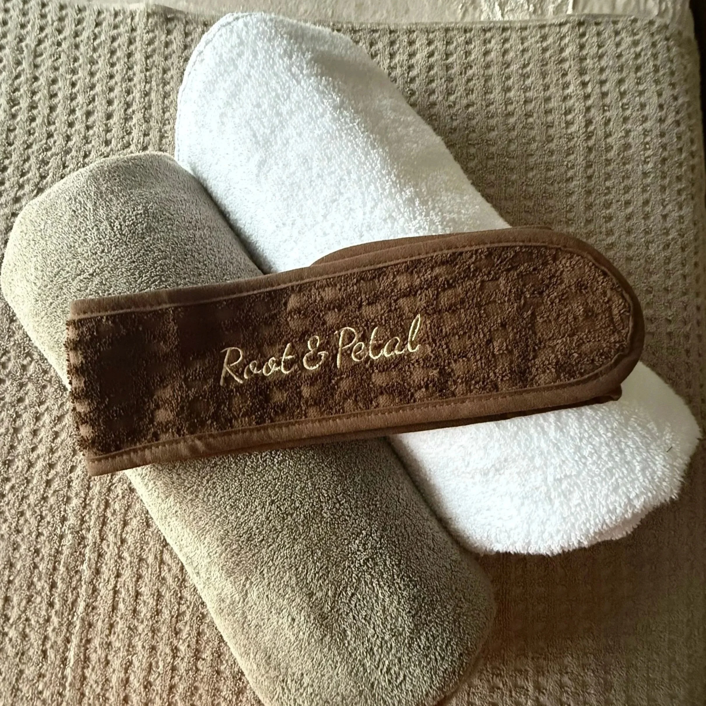 Three rolled towels, two beige and one white, with a brown cloth headband that has 'Root & Pedal' embroidered in cursive, resting on a textured beige surface.