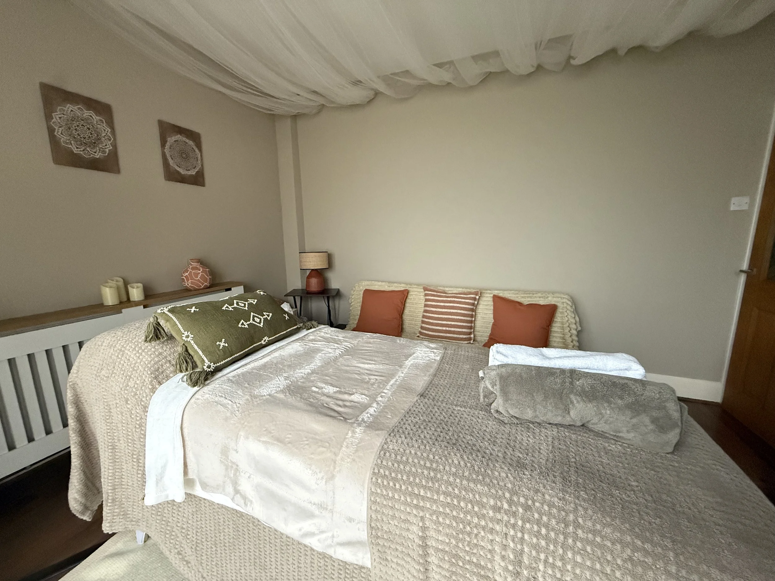 A cozy bedroom with a large bed covered in neutral-toned bedding, multiple pillows, and a nightstand with a lamp. Wall decor includes three framed lace-like patterns, and there are decorative candles and a vase on the dresser.